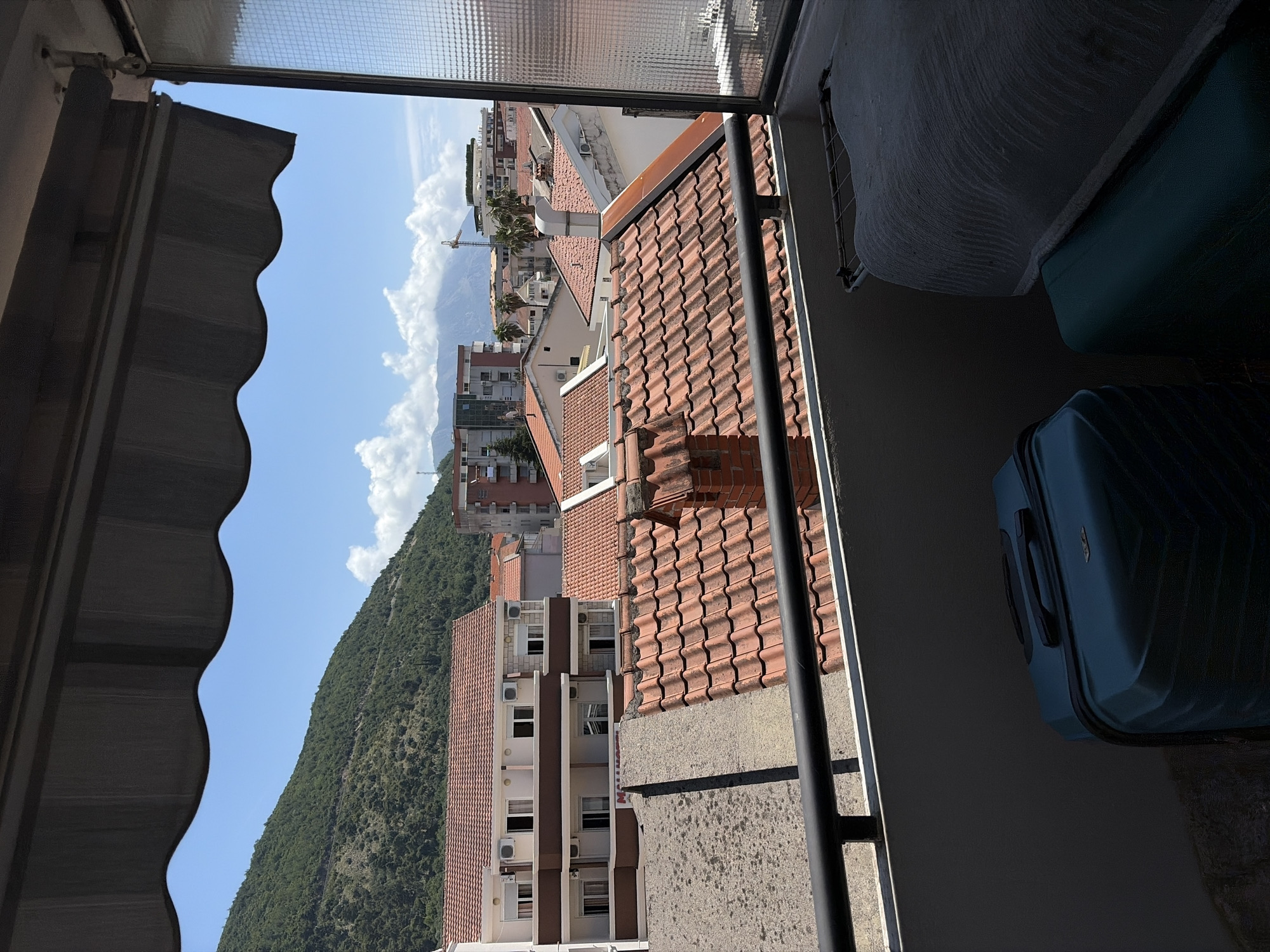 This image captures a vibrant, sunny daytime view looking out from a balcony in Budva, Montenegro. The scene is dominated by a city landscape set against a mountainous backdrop.
In the immediate foreground on the left, a dark grey, scalloped awning is partially visible, indicating a shaded outdoor space. Below it, a grey concrete railing runs across the bottom of the frame, providing a boundary to the balcony from which the photo is taken. On the right, two stacked, dark teal suitcases are partially visible, suggesting storage on the balcony. Above the main opening, a frosted or textured glass panel is seen.
Beyond the balcony, the midground showcases a dense collection of buildings, many featuring characteristic reddish-orange terracotta tiled roofs. Several multi-story residential or commercial buildings are visible, some with white air conditioning units mounted on their facades. A construction crane can be spotted in the distance amongst the buildings, hinting at urban development.
The background is dominated by a large, verdant mountain, thickly covered in green foliage, rising majestically on the left. The sky above is a brilliant clear blue, dotted with white, fluffy cumulus clouds, particularly clustered around the mountain peaks. The bright sunlight casts distinct shadows, enhancing the textures of the roofs and buildings.
There is no discernible activity taking place, and no visible text can be clearly read on any signs or objects. The overall impression is one of a warm, bright day in a coastal city.