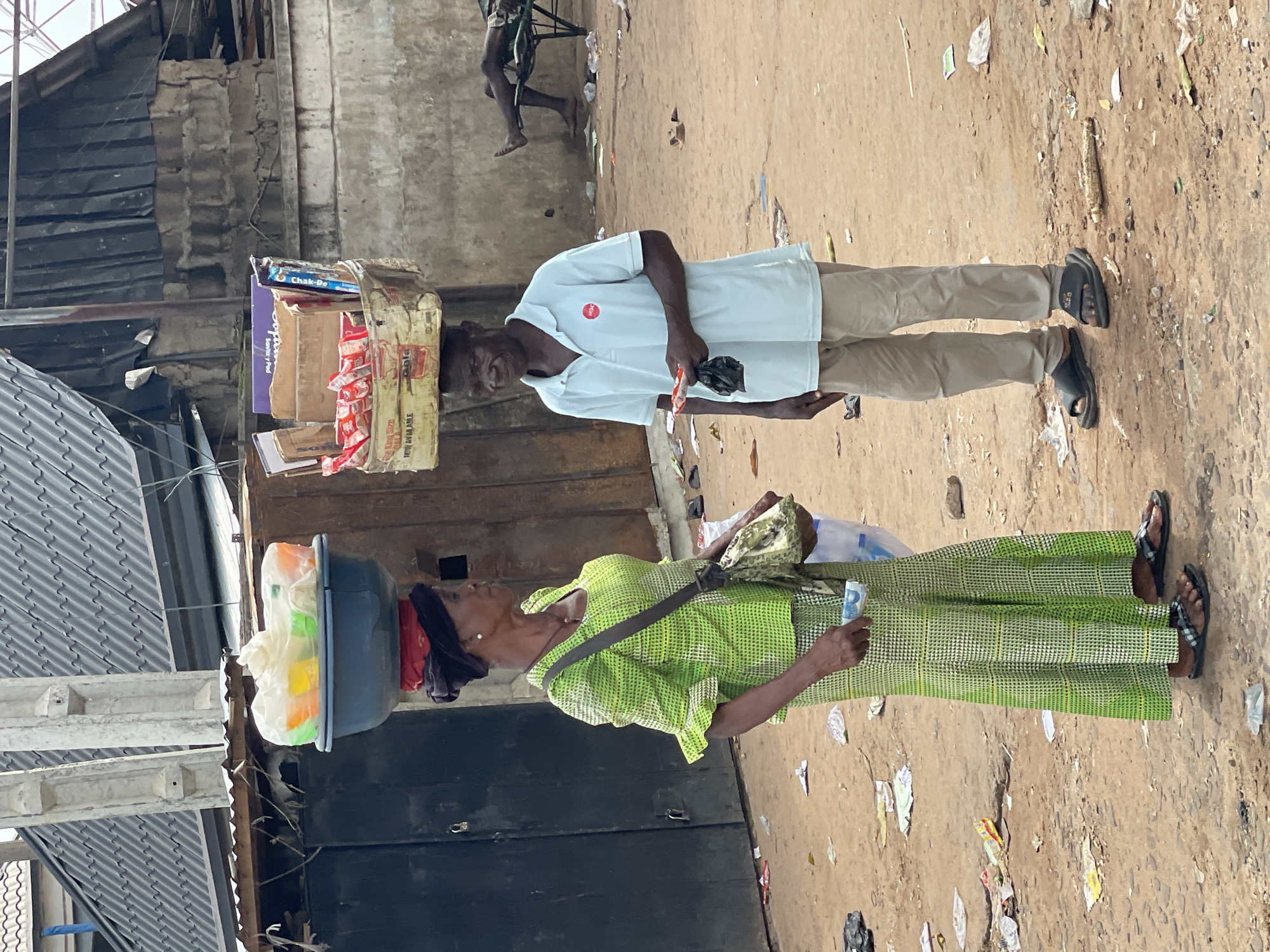 In a street scene in Onitsha, Nigeria, two individuals are prominently featured. A woman in a green patterned dress carries a large, dark container filled with items on her head. She holds a bill in her hand. Adjacent to her, a man in a white shirt and khaki pants balances a stack of goods on his head. The ground is littered with debris and trash. The backdrop consists of buildings with corrugated metal roofing and weathered walls. A person sitting in a chair is visible in the upper background.