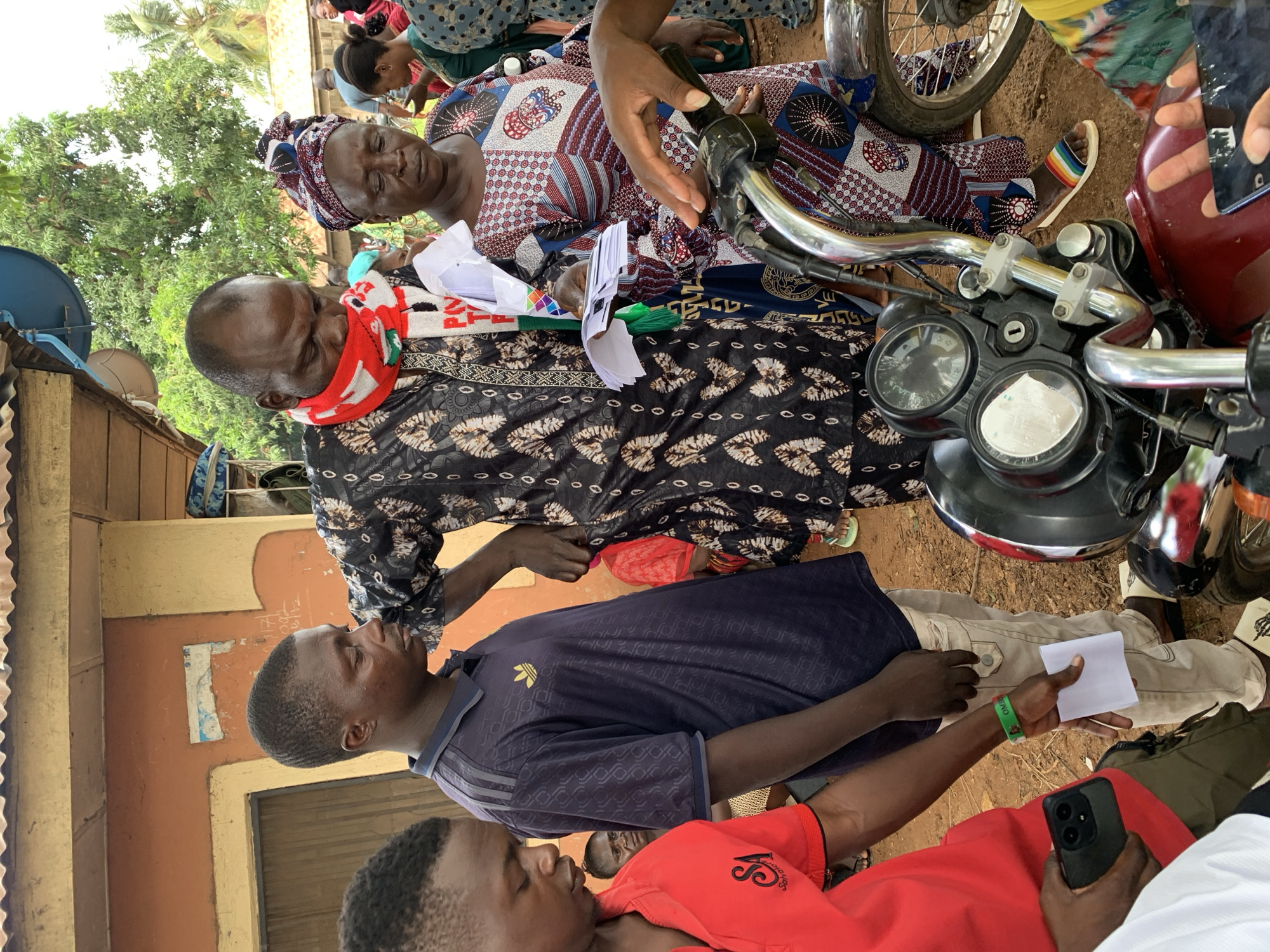 The image appears to have been taken in Akwanga, Nigeria, and shows several people interacting in an outdoor setting. An older man wearing a patterned gown and a red and white scarf around his neck stands near an older woman seated on what looks like a motorcycle. He is holding papers. A younger man in a dark blue shirt stands nearby, also holding papers. Another younger man in a red shirt is seated, holding a smartphone. The ground is dirt, and there is a building visible in the background with a satellite dish.