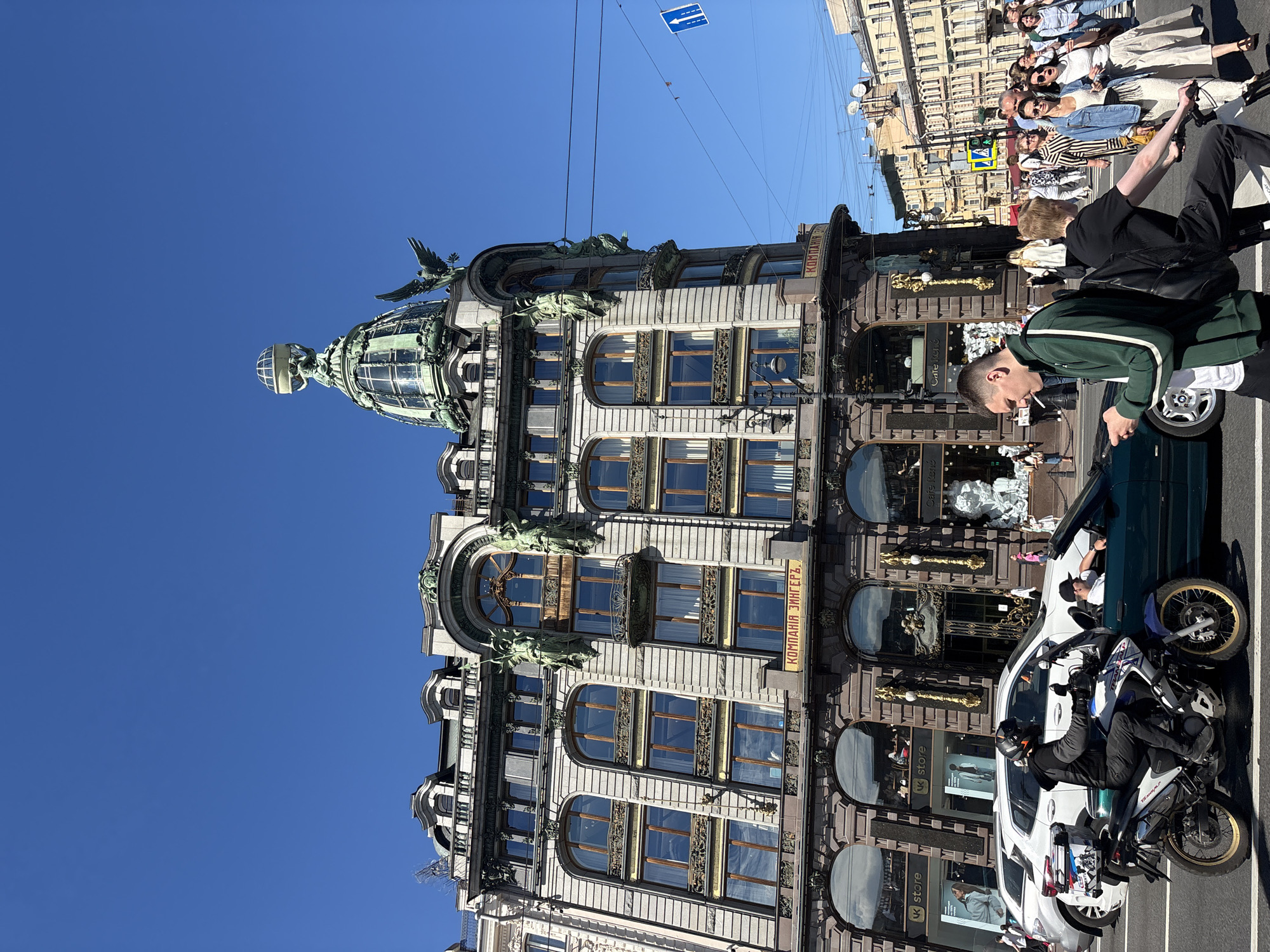 This is a daytime, eye-level, wide shot capturing the Singer House (Dom Knigi) located on Nevsky Prospekt in Saint Petersburg, Russia. The iconic building, with its distinctive green dome topped by an eagle statue and a glass globe, dominates the upper half of the frame against a clear blue sky. The facade features ornate architectural details, numerous windows, and decorative sculptures. Below the building, the street scene unfolds with activity. A motorcycle carrying a rider in a helmet and dark clothing is in the foreground, positioned on the left side of the frame. A dark green car is to its right, with a person visible inside. A man in a green tracksuit and a black backpack is walking in the mid-ground, looking down at his phone and holding a cigarette. A crowd of pedestrians, dressed in casual summer attire, are walking on the sidewalk and crossing the street, with some carrying shopping bags. Traffic signs and overhead wires are visible, indicating a busy urban intersection. The lower portion of the buildings across the street shows more shops and businesses. The overall atmosphere is lively and bright, under a sunny sky.