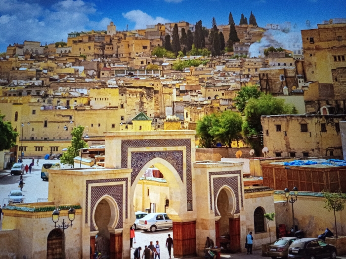 A vibrant cityscape unfolds under a blue sky with scattered clouds. Multi-story buildings in earth tones cascade down a hillside, interspersed with trees. An ornate gateway dominates the foreground, featuring intricate tilework and arched entrances, with people passing through. Cars are parked on the side, and streetlights add to the urban scene.