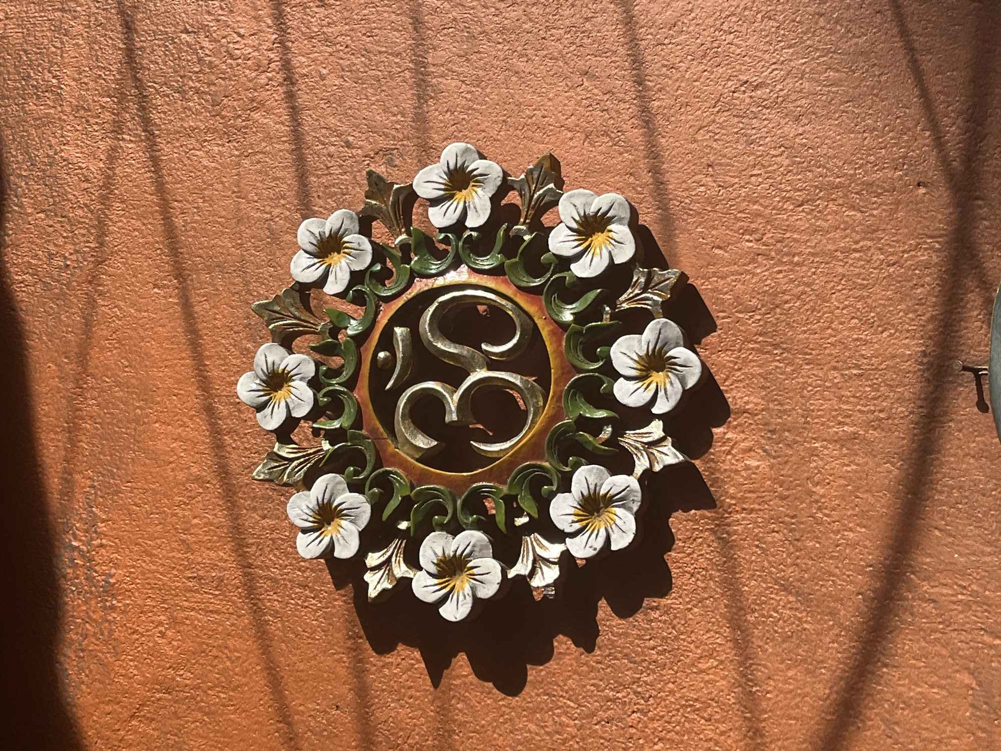 A medium, eye-level shot shows a light-bronze, textured wall in Ubud District, Indonesia, upon which is hung a circular, decorative metal piece.
The metalwork is comprised of an ornate gold center containing a silver “S” shape, surrounded by a ring of red-gold paint. Beyond this ring is a green, plant-like form interwoven with 10 evenly-spaced, white-petaled flowers with yellow centers. Gold leaves protrude from between each flower. The shadows from above create a light-and-dark vertical striping along the wall.