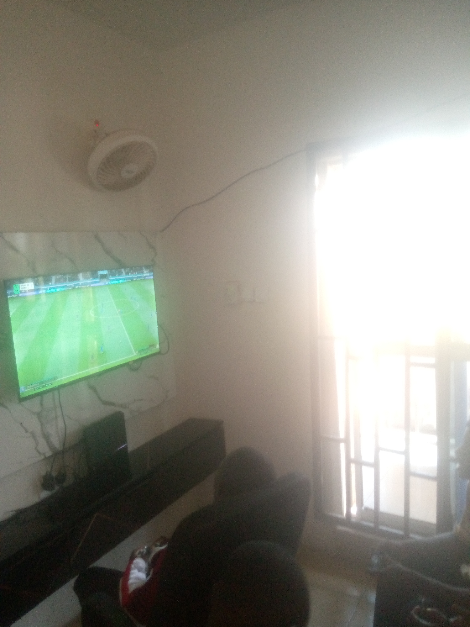 The image depicts an indoor entertainment space, likely a living room, located in Jalingo, Nigeria. The scene is brightly illuminated by an intense natural light source originating from a large, gridded window on the right side, which is significantly overexposed. Additional light emanates from a television screen mounted on the left wall.
On the left wall, a flat-screen television displays a video game or live match featuring a green soccer (football) field. Below the television, a dark, elongated entertainment console or shelf holds a dark, rectangular electronic device, possibly a gaming console, with associated cabling visible. The wall behind the television exhibits a subtle, light-toned marble-like pattern. Above the television, a white wall-mounted fan with a red indicator light is installed. A black electrical cable runs horizontally across the upper portion of the left wall and extends diagonally towards the right.
On the right side of the room, the large window with multiple vertical and horizontal divisions allows strong sunlight to penetrate, causing significant glare. To the left of the window, on a light-colored wall, a white electrical outlet plate with two discernible switches or sockets is mounted.
At least three individuals are present, seated in dark-colored, upholstered furniture in the foreground and midground. In the lower foreground, a person is seated with their back partially to the camera, wearing a dark top over a red long-sleeved garment, and holding a white object consistent with a video game controller. Another person is partially visible in the mid-right, seated near the window. A third individual's back is visible in the lower right corner of the frame. The individuals appear to be engaged in watching or playing the content displayed on the television.