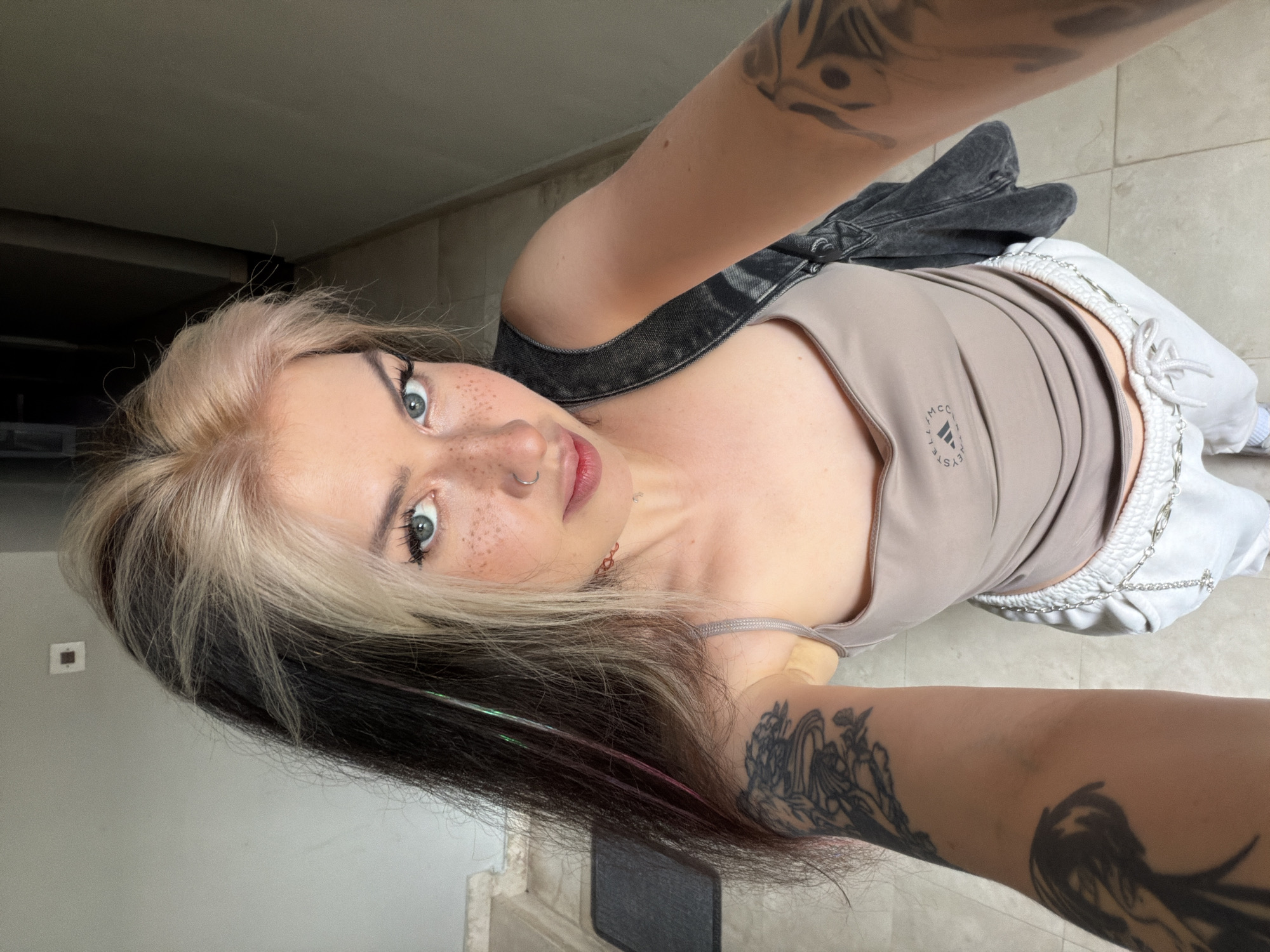 A young woman with dyed hair is taking a selfie. She is wearing a light brown tank top, white sweatpants with a chain detail, and a dark denim jacket draped over her shoulder. The woman has tattoos on both arms and a nose ring. She is looking directly at the camera with a slight smile. The background appears to be an interior space with light-colored tiled floors and a gray wall. There are no indicators of the location in Budva, Montenegro.