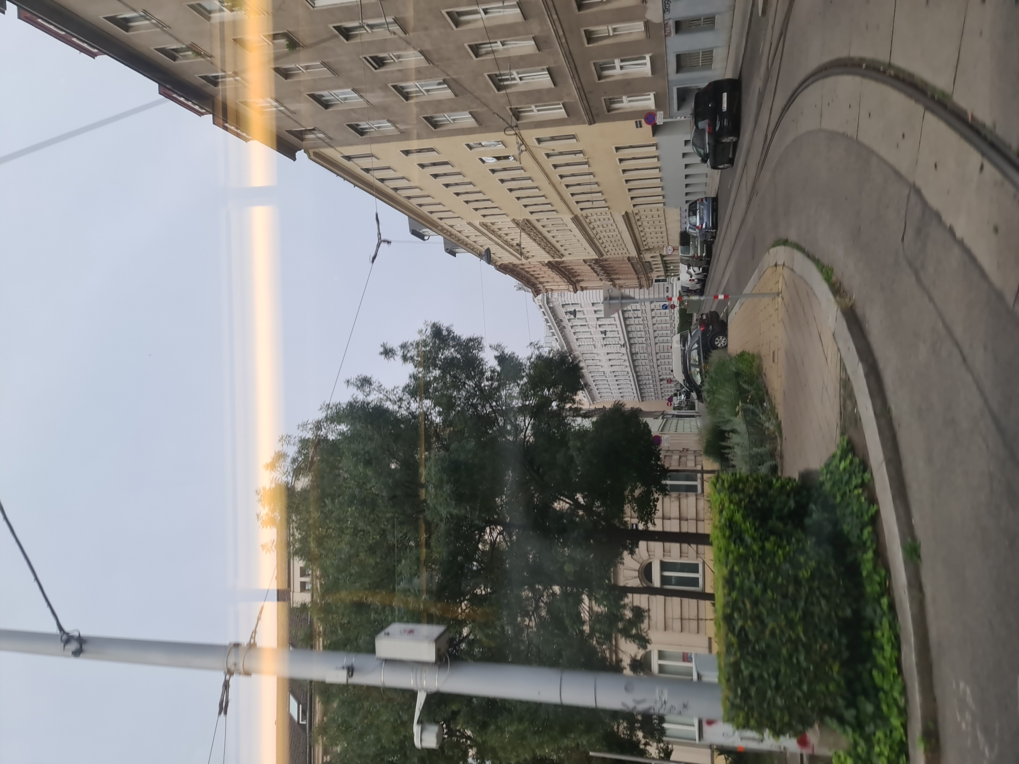 The image captures a street scene in Vienna, Austria, likely taken from a moving tram. The perspective shows a curved street with tram tracks running through it. On the right side of the street are several apartment buildings with neutral-colored facades, multiple windows, and some architectural details like cornices. A dark-colored car is parked on the right side of the street. Further down the street, more buildings are visible, and a row of cars is parked along the left side. The left side of the image features a lamppost with a white rectangular box attached to it, a lush green hedge, and trees with dense foliage. The sky is overcast with a pale, diffused light, suggesting it is daytime. Reflections are visible on the surface from which the image is taken, possibly a tram window.