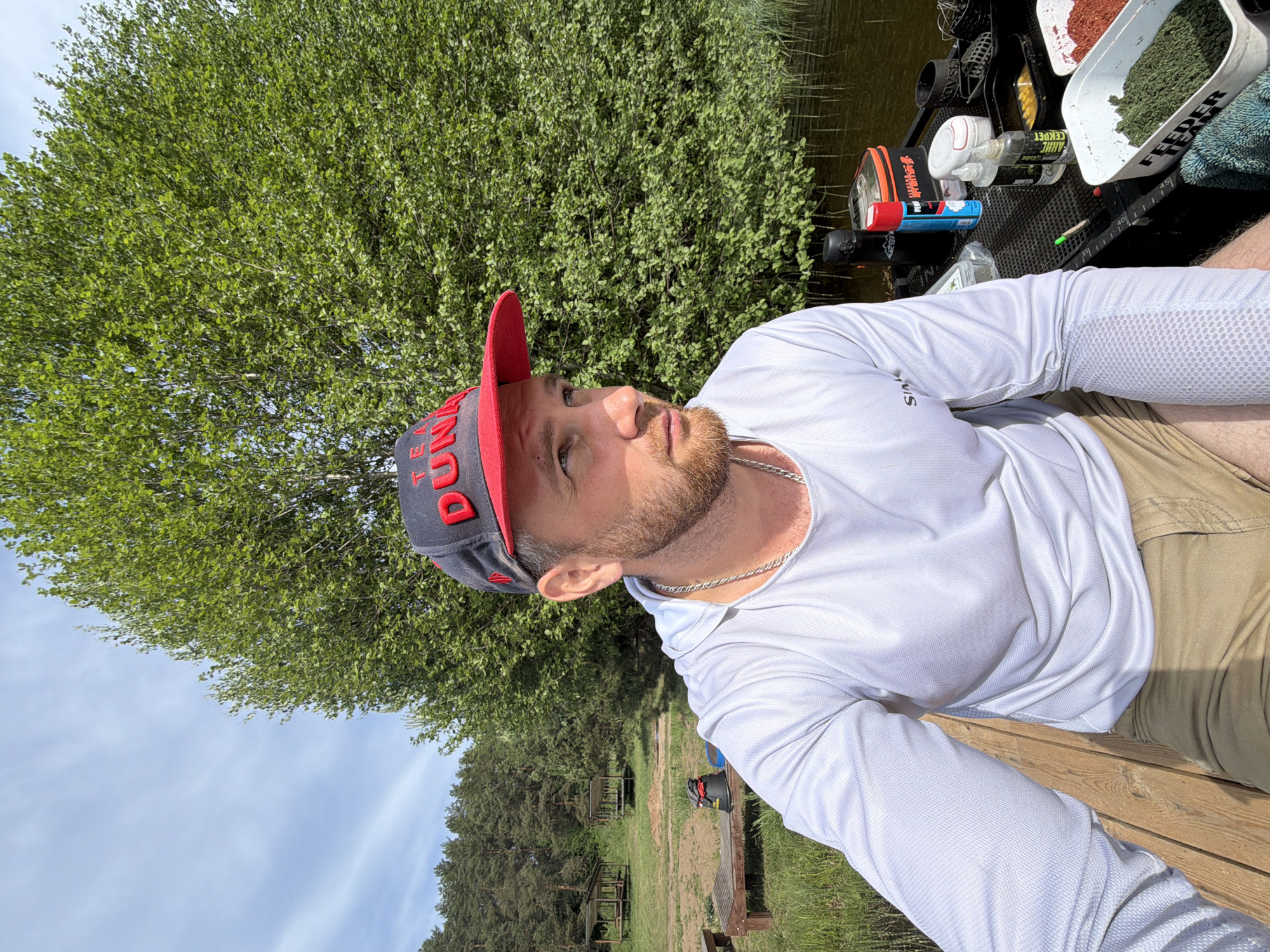 A man with a beard is sitting at the edge of a pond near Prikamskii, Russia, wearing a white long-sleeved shirt, khaki pants, and a baseball cap with a red bill. He is looking upward and to the side. Behind him is a dense green tree. To the man's right is fishing equipment, including containers of bait. In the background, there are picnic tables and more trees, suggesting a park area. The sky is partly cloudy.