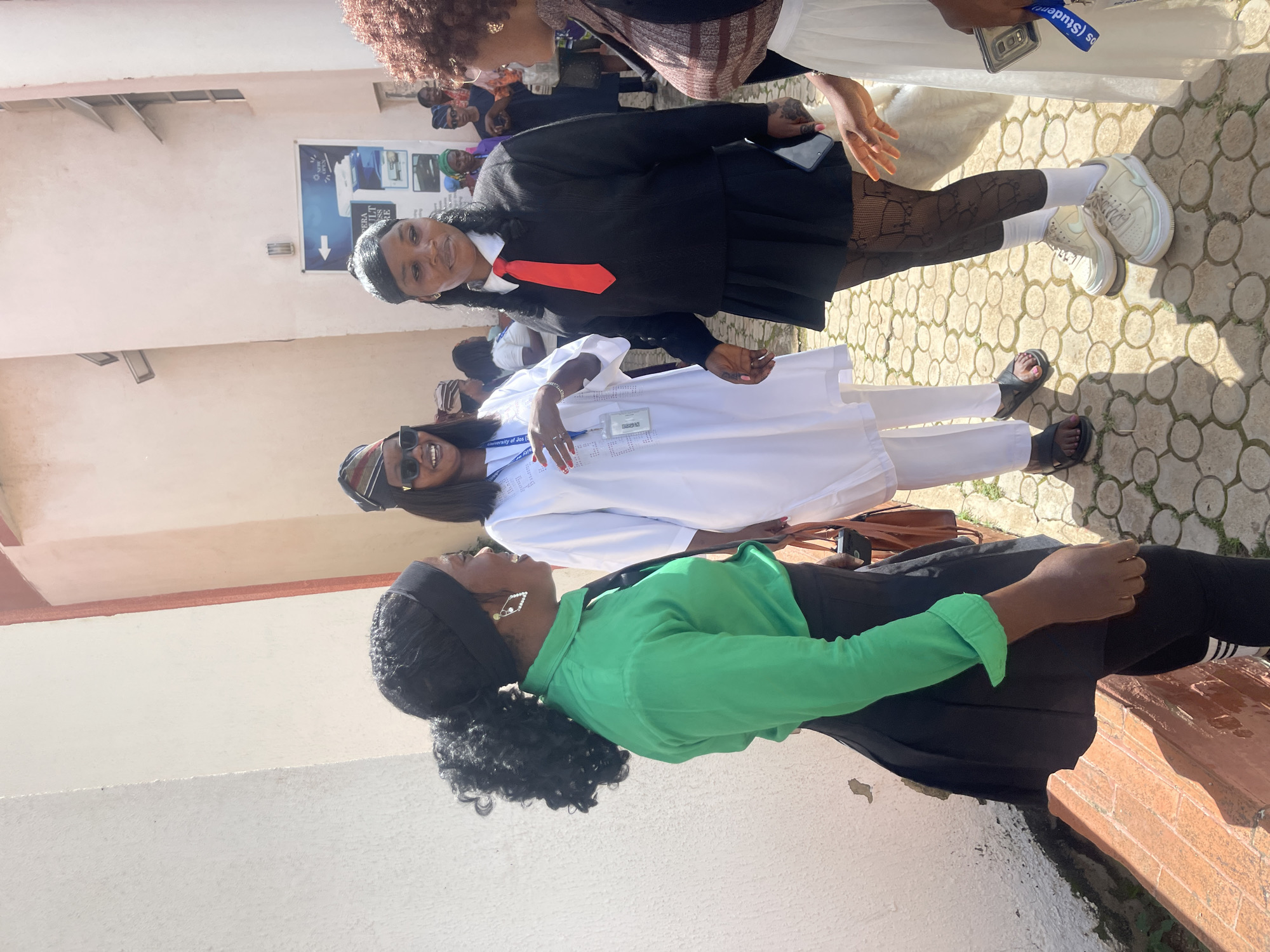In Jos, Nigeria, four young women, appearing to be students, are gathered outdoors. The foreground is occupied by two women. One, on the left, wears a green long-sleeved shirt, a black mini-skirt, black leggings, and a black headscarf. She is looking to her right, and her mouth is open as if speaking or laughing. The second woman in the foreground, on the right, is dressed in a black sweater with a white collar and a red tie, a black mini-skirt, and patterned black stockings with white socks and cream-colored platform sneakers. She is looking to her left and her mouth is partially open. To their left, another woman in a white outfit with a patterned trim and a matching headscarf is smiling and looking to her left. In the background, several other individuals, some wearing headscarves, are visible, suggesting a larger gathering. The environment appears to be a courtyard or walkway with textured paving stones and a light-colored building wall with an open doorway and a sign in the background. The lighting suggests it is daytime.