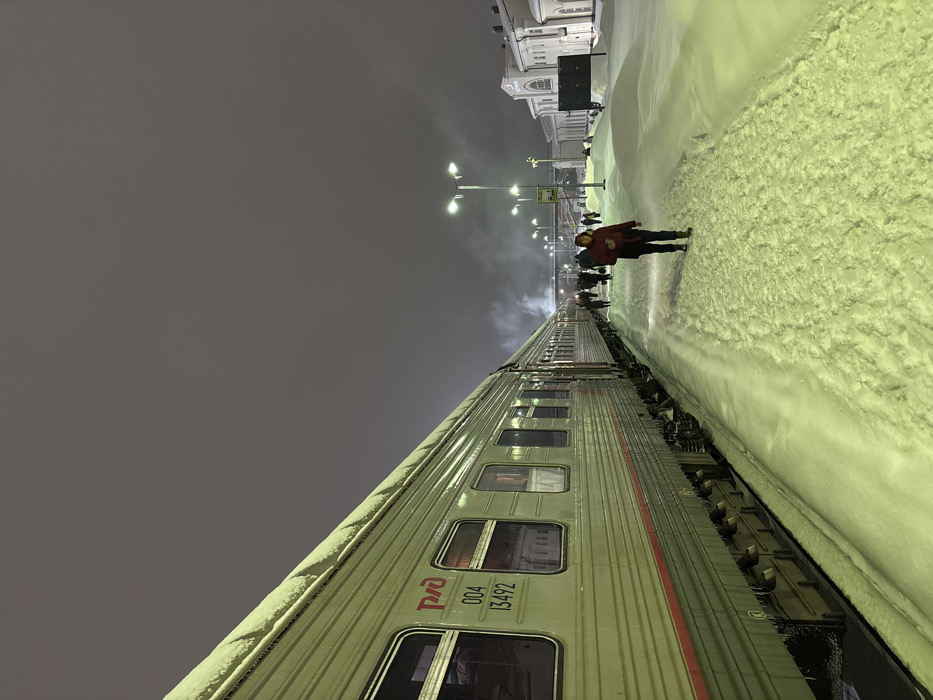 A train stretches into the distance under an overcast sky and artificial lights. The train cars are silver with dark-framed windows and a red logo on the side. A thick layer of snow covers the ground alongside the train, forming a high bank. A person in a red jacket stands on the snowy ground near other figures, with a building in the far background.