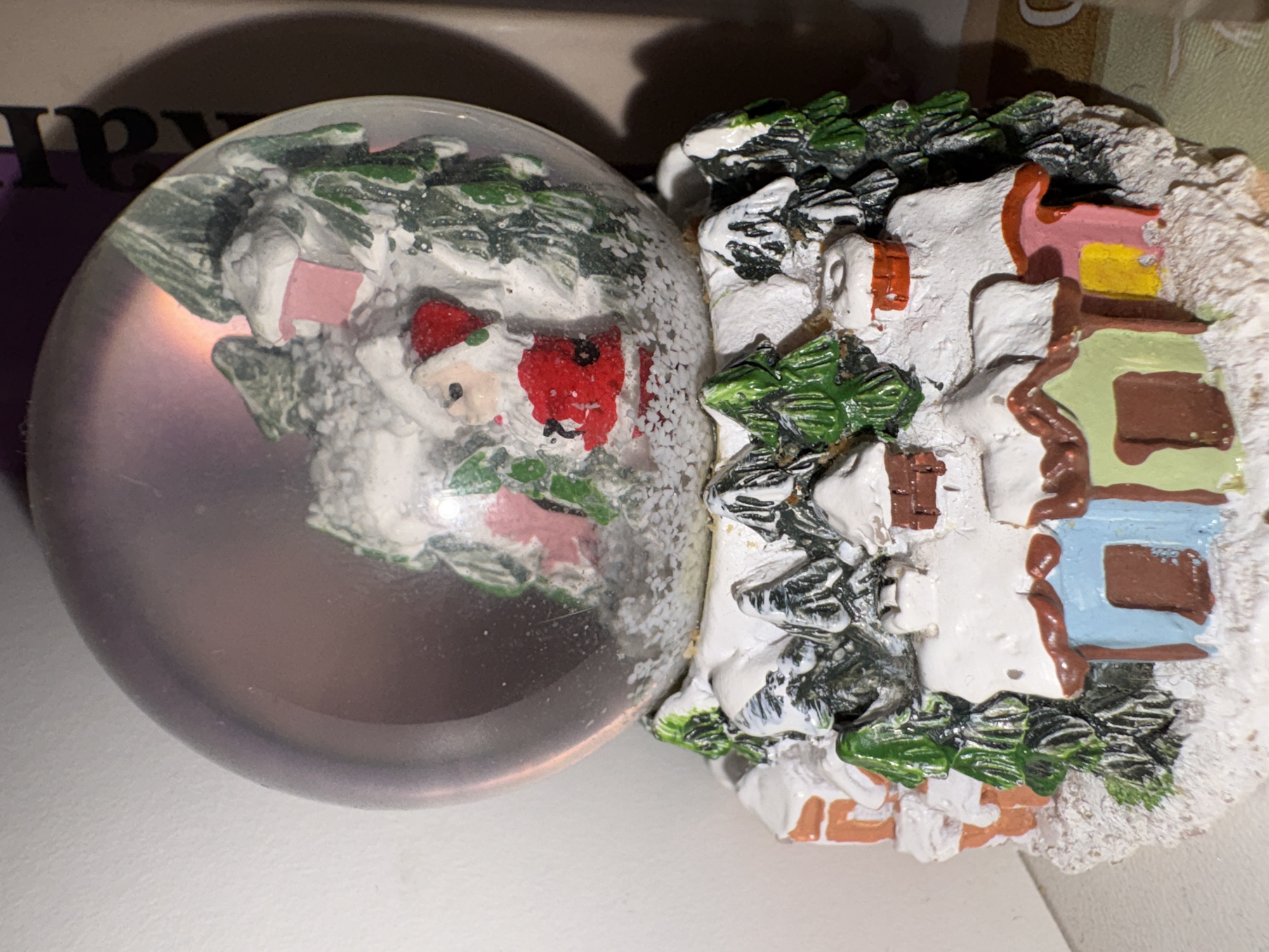 A close-up captures two decorative Christmas ornaments resting on a white surface. On the left, a spherical snow globe encases a miniature scene of Santa Claus surrounded by snow-covered evergreens and a small pink building. The snow globe is transparent, revealing the interior scene and creating light refraction. To the right of the snow globe is a decorative house ornament with a snowy roof and colorful accents. The house is adorned with green evergreen details. The background is partially visible and indistinct due to the close proximity of the shot.
I am unable to provide information about Izhevsk, Russia, or any location information not visibly present in the image.