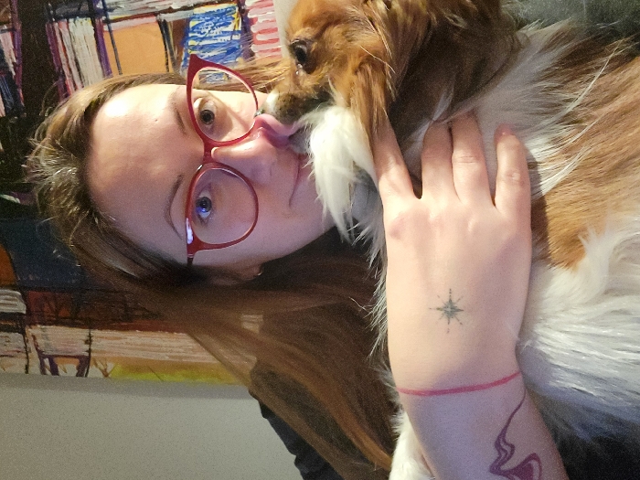A woman with red-framed glasses is seen in a close-up selfie, being licked on the face by a brown and white Cavalier King Charles Spaniel. The woman's left hand is resting on the dog's back. A compass tattoo is visible on her hand, along with a red string bracelet. A corner of a colorful, abstract painting is visible in the background.