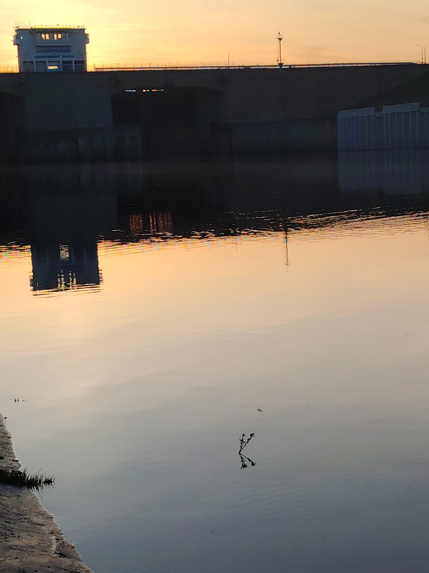 The image captures a sunset view of the Kama Hydroelectric Station in Chaikovsky, Russia. The hydroelectric station, a massive concrete structure, dominates the horizon with a control building on top. A series of arches and support structures are visible beneath the bridge. The station's reflection is cast across the water surface, slightly distorted by ripples. In the foreground, a small, green plant or twig floats near the water's edge. The sky is a mix of orange and yellow.