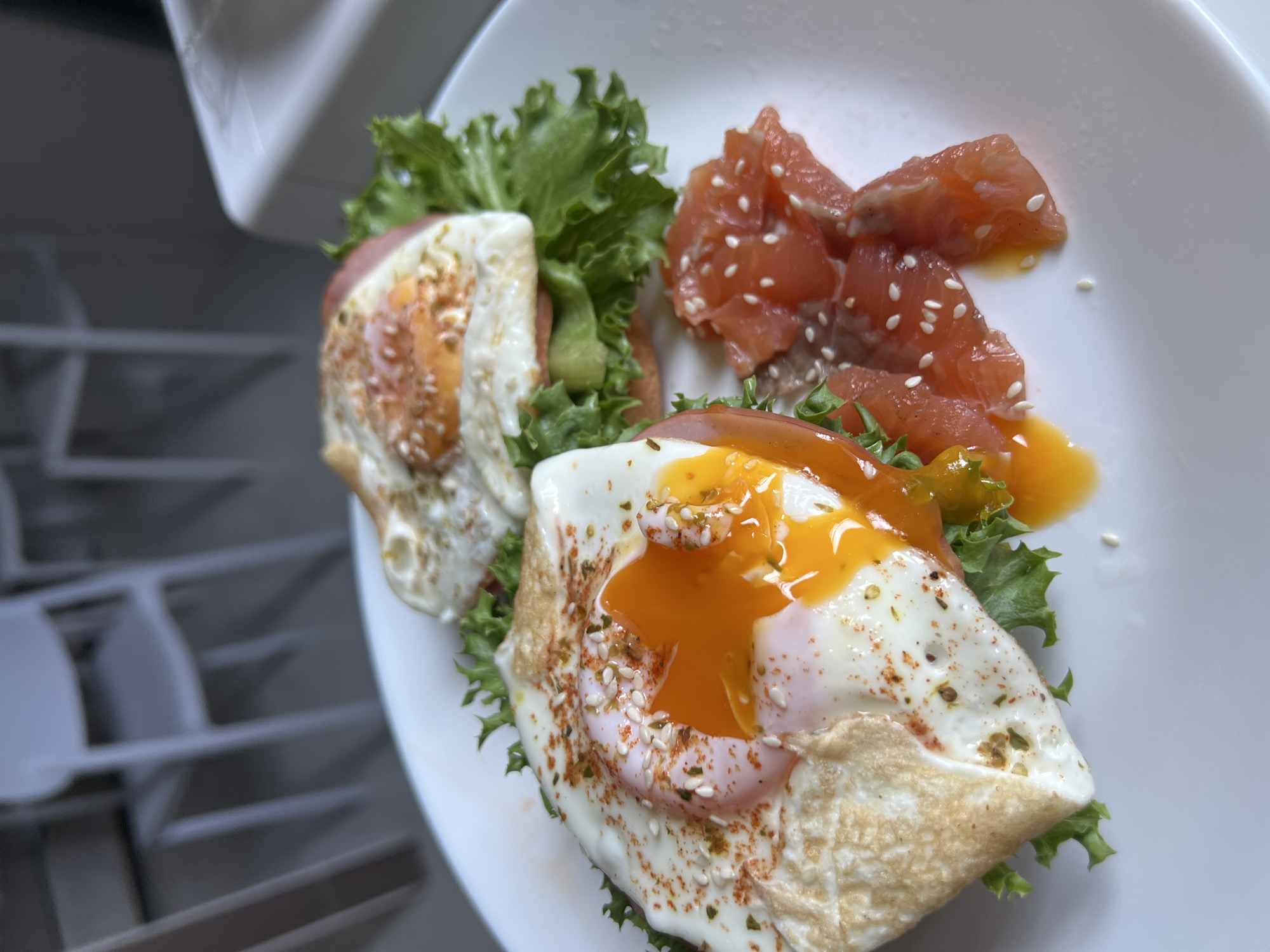 A close-up, high-angle shot shows a white plate with a breakfast dish. The dish consists of two open-faced sandwiches on a bed of lettuce. Each sandwich has a fried egg on top, with the yolk of the left egg runny and spilling onto the plate. Both eggs are sprinkled with seasonings and sesame seeds. To the right of the sandwiches are slices of raw salmon, also sprinkled with sesame seeds. The background is blurred and shows white furniture and a white surface, possibly a table. The scene appears to be indoors, in a bright, well-lit environment. No people are visible in the image.