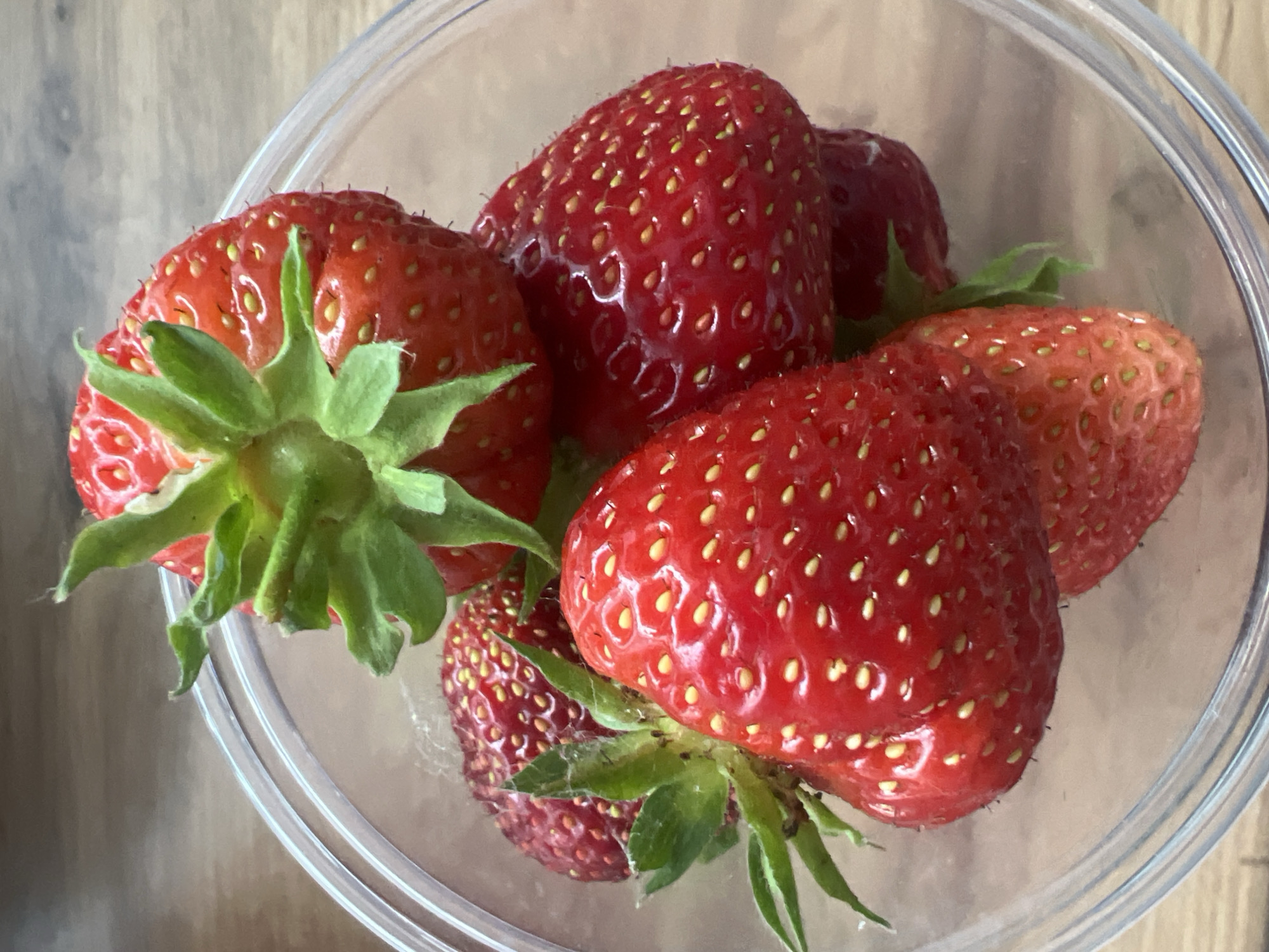 This is a close-up, overhead shot of several ripe strawberries in a clear plastic container. The strawberries are a vibrant red color with visible seeds dotting their surface. The green leafy tops of some of the strawberries are prominent, especially one on the left side of the frame. The container is transparent and the light reflects off its surface, creating bright highlights. The background consists of a wooden surface, likely a table. There are no people, text, or specific time-of-day or weather cues visible. The setting appears to be domestic, with the strawberries likely prepared for consumption.