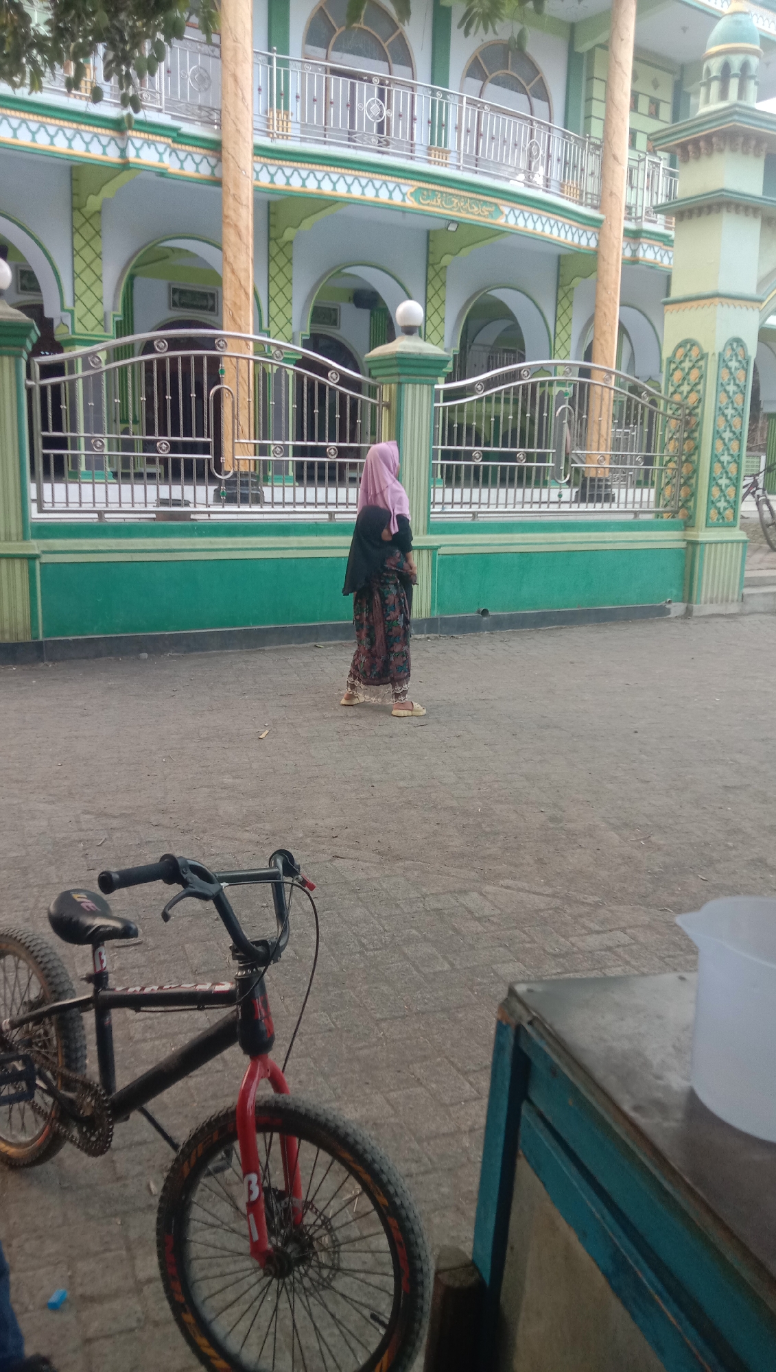 A side view of a person wearing a pink hijab and a patterned dress is captured walking on a paved outdoor area in front of a mosque. The mosque features green and white accents, with ornate metal railings and arched windows visible. A black bicycle with red accents is parked in the foreground on the left, and a turquoise-colored wooden structure with a white plastic container on top is visible on the right. The scene appears to be in Ngempit, Indonesia.