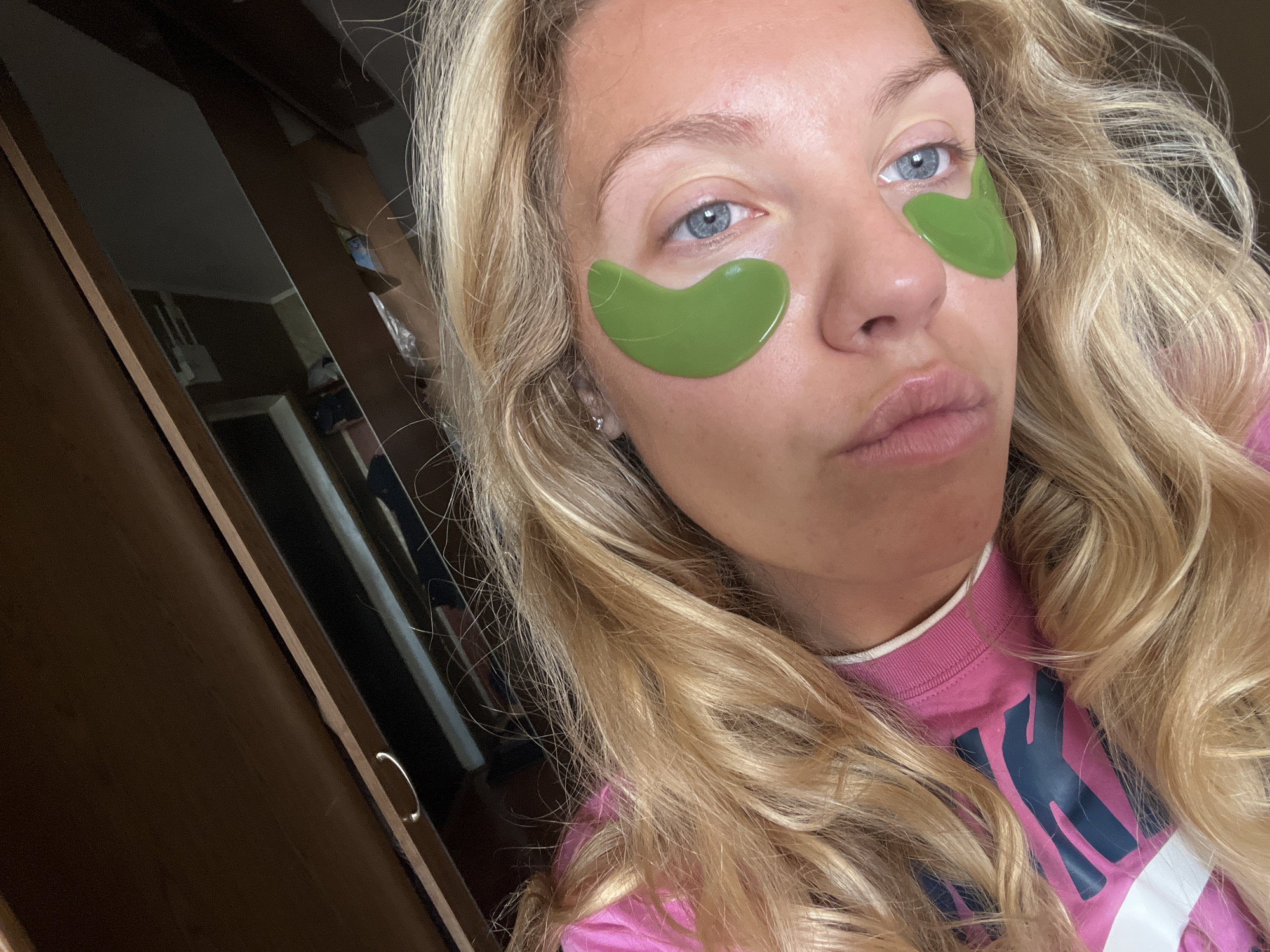 A young Caucasian woman with blonde, wavy hair is lying down, looking up at the camera. She is wearing a pink t-shirt with a dark blue logo. Green under-eye patches are applied to her skin. She has light blue eyes and her lips are slightly pursed. The background shows a reflection in a mirrored surface, possibly a closet door, revealing a dimly lit room with furniture and what appears to be a doorway. The image does not contain enough information to determine a specific location in Khimki, Russia.
