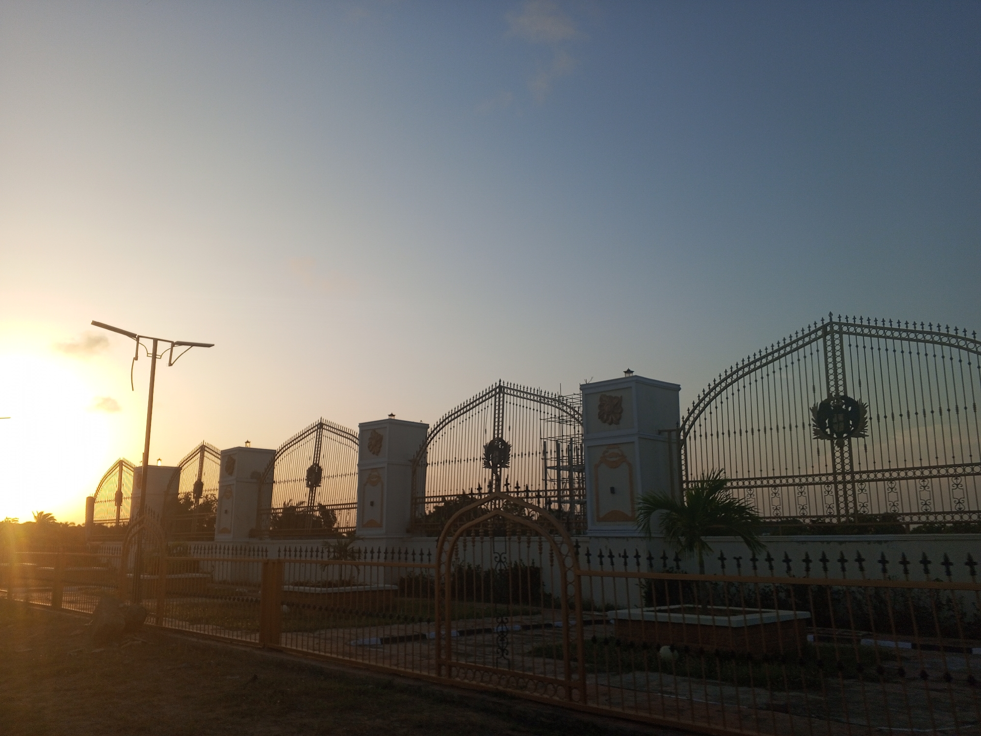 A low-angle, medium shot depicts a series of ornate, arched gates stretching into the distance under a sky gradient from yellow-orange near the horizon to pale blue overhead. The gates are constructed from dark metal, with a decorative flourish at the top of each. They are interspersed with squat, cream-colored pillars adorned with golden-yellow embellishments. Behind the gates, green foliage is visible. A lower fence of golden-yellow metal runs along the foreground, obscuring the view of what appears to be a landscaped area. To the left, a utility pole rises against the bright sun. Scaffolding is visible behind one of the pillars, suggesting ongoing construction or maintenance. The scene is washed with warm light, indicating sunrise or sunset. Based on the prompt, the location is Tawpo, Nigeria.