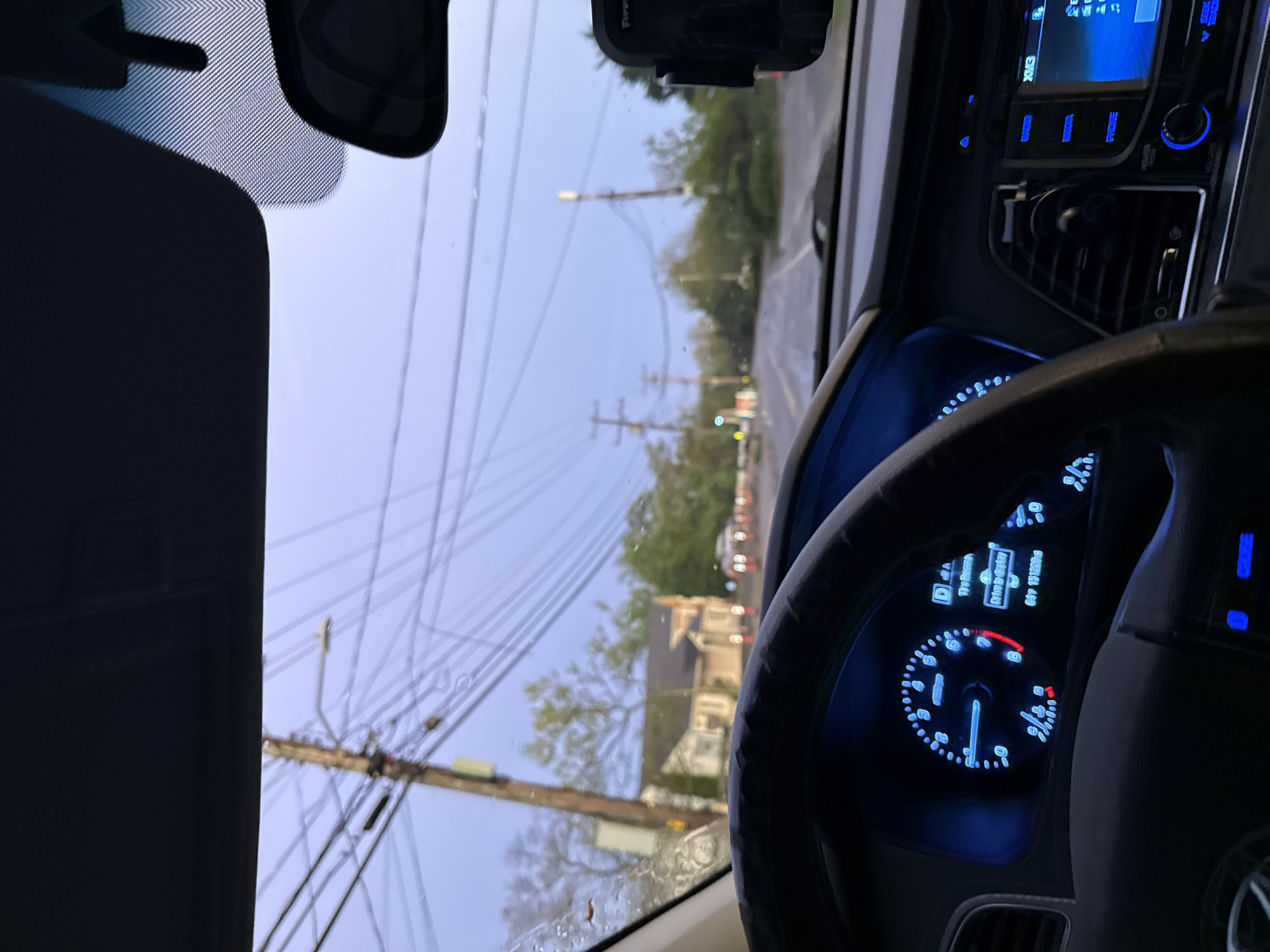 The image shows the view from inside a car, likely in motion. The steering wheel and dashboard are prominent, with illuminated gauges. Outside, telephone poles with numerous wires are visible along a residential street. A house is seen on the right side of the road. Based on the architectural style of the house, the vegetation, and utility poles, the location may be in the United States. Without more information, it is impossible to confirm that the location is Provincetown, United States. The sky is overcast, suggesting it may be early morning or late evening.