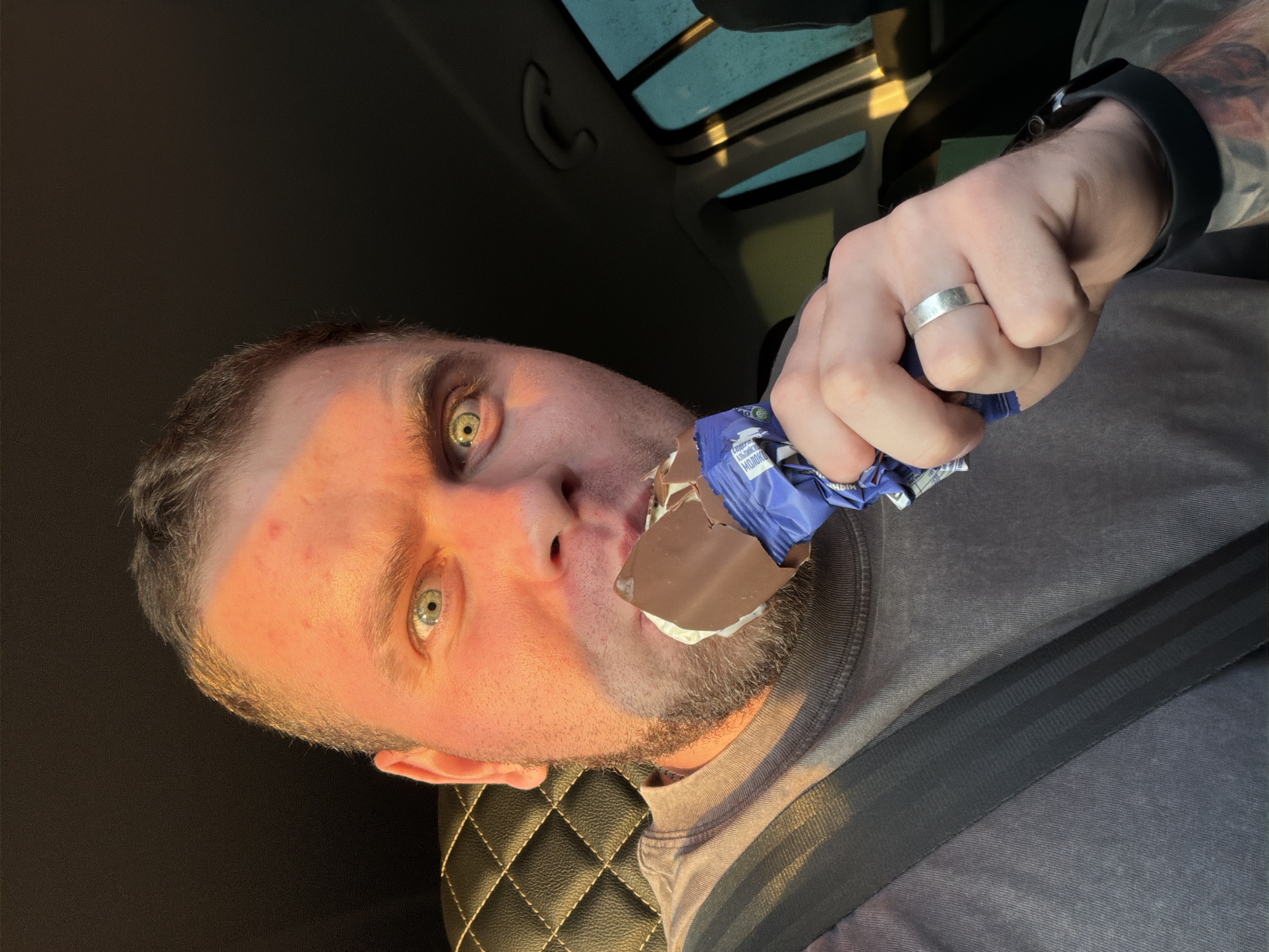 The image is a selfie of a man in a car, eating a chocolate-covered ice cream bar. He is wearing a gray t-shirt and a seatbelt. He has a silver ring on his finger. The interior of the car has black leather seats and a sunroof. The location of the image is not mentioned, nor is the city of Полозово, Russia.