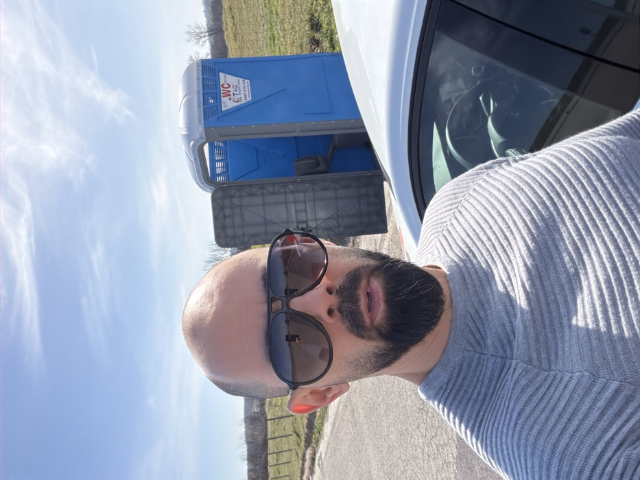 A front-facing image captures a male subject taking a selfie outdoors in Dositeevo, Bulgaria. The man, appearing bald with a dark, full beard and wearing dark aviator-style sunglasses, is dressed in a light grey horizontally ribbed long-sleeved sweater. He stands on an asphalt surface, positioned to the left of a white vehicle, likely a sedan, with its driver's side visible. Behind the vehicle and the man, within a grassy field, a blue and grey portable toilet with an open door is present. The portable toilet bears the inscription "WC T&G" and the phone number "0887 014 416" on its side. A wire fence separates the asphalt surface from the grassy field, which extends to a distant treeline under a bright blue sky with sparse white clouds. The scene is brightly lit by natural sunlight.