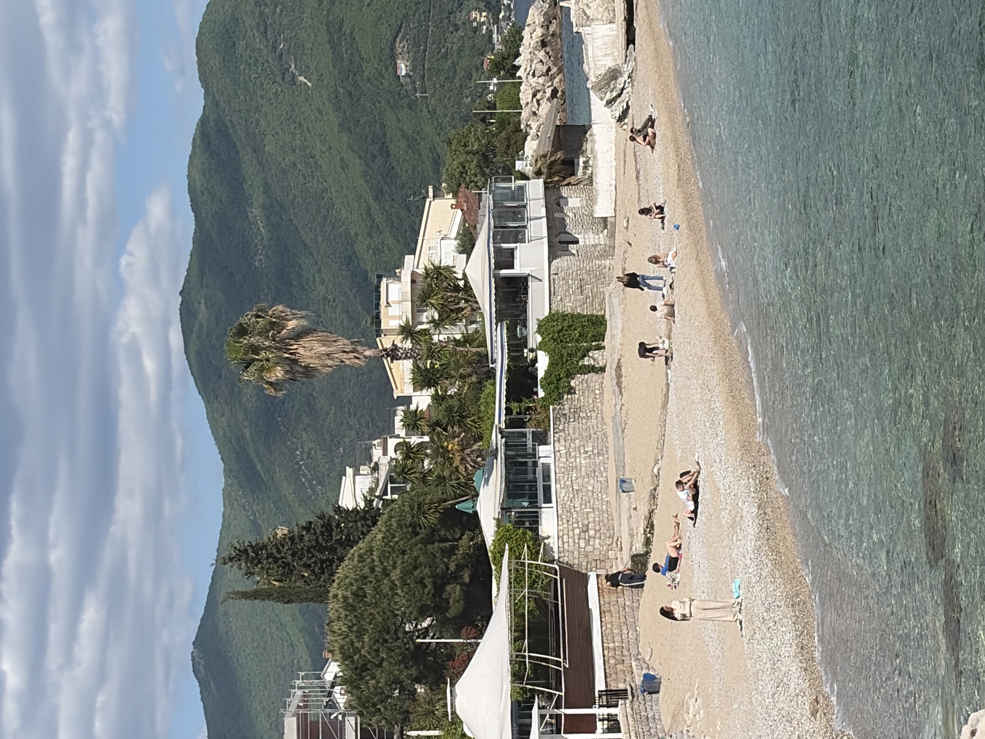 The image presents a vertical panoramic view of a beach near Herceg Novi, Montenegro. A mountain covered in dense green vegetation rises to the left of the frame, with a blue sky visible above. A coastal strip features several buildings and structures with white walls and tiled roofs. Palm trees are interspersed among the buildings. The beach itself has a sandy surface and features numerous people engaged in various activities, such as sunbathing, walking, and sitting. The water is clear and turquoise-colored.