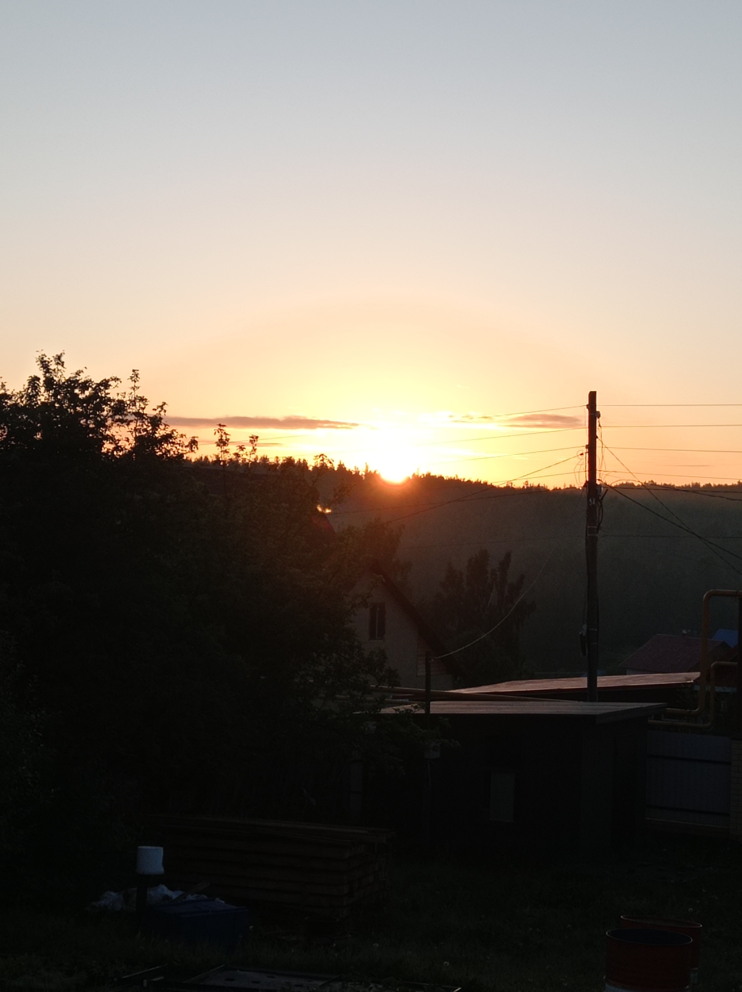 The image depicts a silhouetted rural landscape at sunset. The sky is a gradient of pale blue to soft orange and yellow as the sun sets behind a distant treeline. In the foreground, dark trees and foliage dominate the left side of the frame. A wooden structure with a flat roof and a small window is visible in the mid-ground. To the right of this structure, a utility pole with wires stands tall, and beyond it, parts of other buildings and a fence can be seen. The ground is covered in grass and in the lower left corner, there are some stacked wooden planks and a white bucket. Two red barrels are visible in the lower right corner. The scene suggests a quiet, possibly evening setting in a rural area near Degtyarsk, Russia, with residential buildings and infrastructure elements present. There are no people or active interactions depicted.