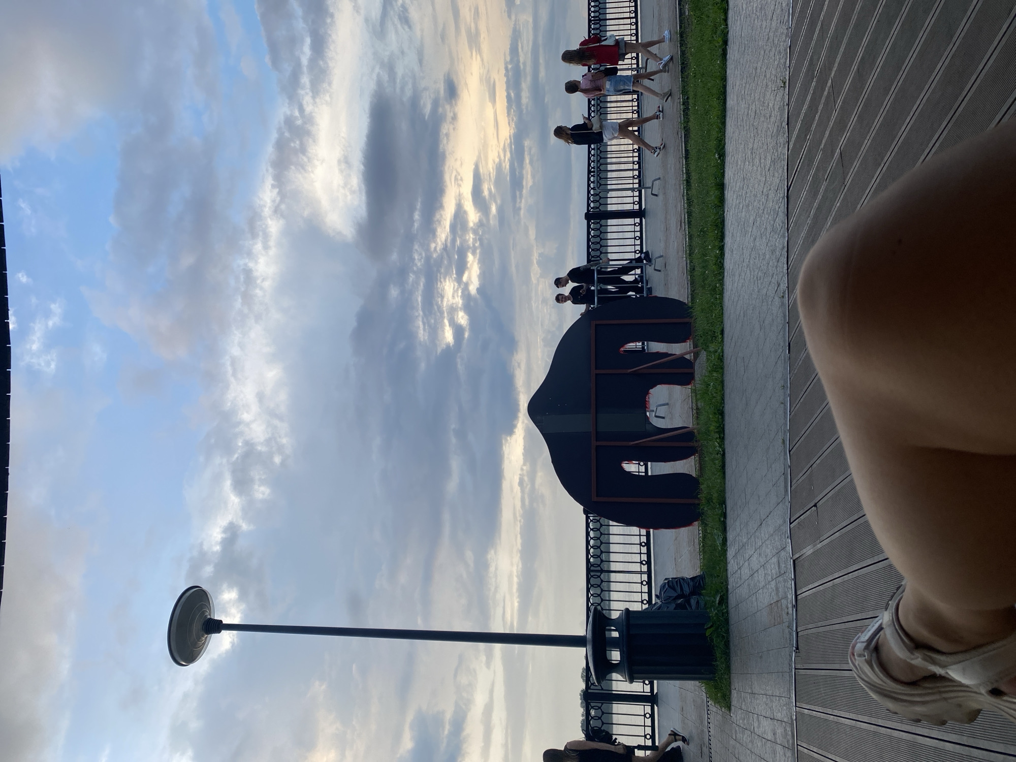 The image shows a cloudy sky above a waterfront promenade in the city of Perm, Russia. A lamppost stands on the left side of the image, next to a trash can. A black, tent-like structure with a cutout opening is visible in the middle ground, situated on the promenade. Several people are walking along the promenade. In the foreground, a person's legs and feet are visible, resting on a wooden deck. The scene appears to be a public park or recreational area along the Kama River.