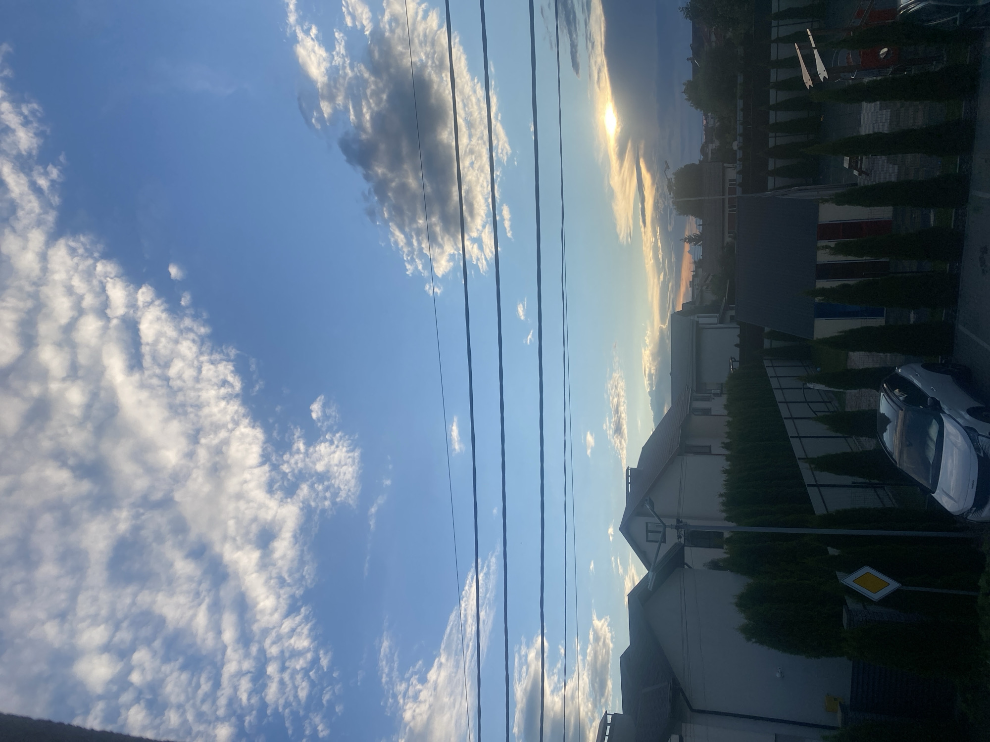 The image depicts a residential area in Novi Petrivtsi, Ukraine, likely during the late afternoon or early evening, as indicated by the low sun and the sky's colors. The scene is dominated by the sky, which features a mix of blue and scattered white clouds. Four parallel power lines traverse the midsection of the frame horizontally. Below the sky and power lines, the tops of several modern-looking houses with grey and white facades are visible, interspersed with rows of tall, dark green coniferous trees. A black metal fence runs along the property lines. In the foreground, a white SUV is parked, partially obscured by the fence and trees. To the left of the SUV, a yellow diamond-shaped road sign is mounted on a post. Further to the right, a small, brightly colored shed or playhouse with red, white, and blue panels is visible, along with a children's playset. The overall impression is one of a quiet suburban setting under a dramatic sky.