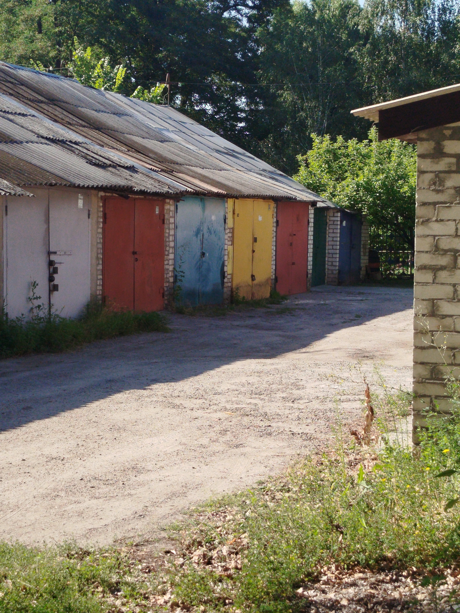 The image depicts a row of garages with corrugated metal roofs and various colored doors, including white, red, blue, and yellow. The garages are constructed of brick. In the foreground, a dirt road with tire tracks leads towards the garages, casting a shadow from an unseen object. On the right side of the image, a partially constructed brick building with exposed mortar is visible. Green foliage and some dry leaves are present around the garages and in the foreground. The background features a dense canopy of green trees. The scene appears to be in an outdoor urban or semi-urban setting.
