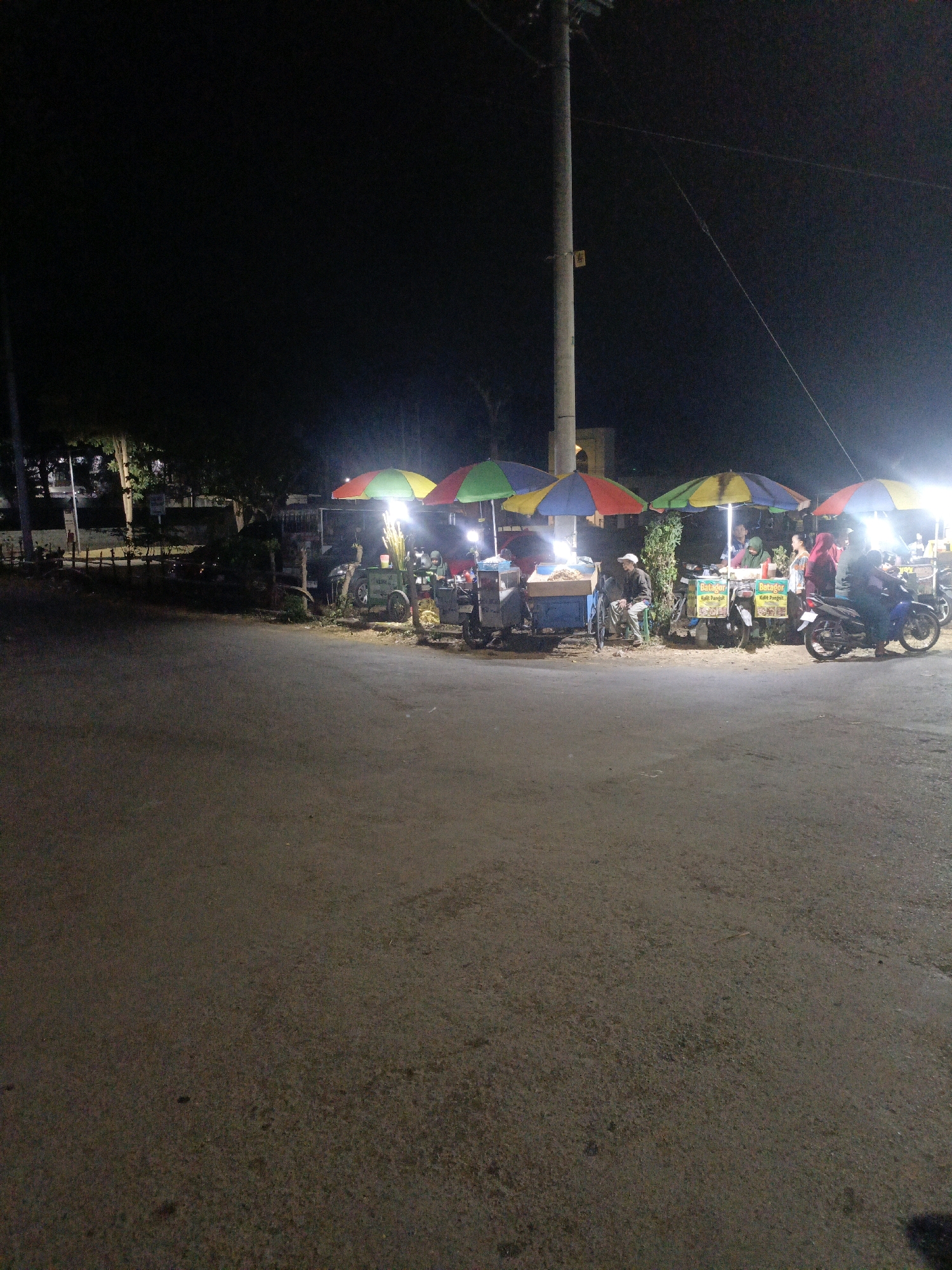The image depicts a street scene at night in Temenggungan, Indonesia, likely near a food stall area. In the foreground, a paved road is visible. In the background, several food carts are set up under colorful umbrellas, illuminated by bright lights. People are interacting around the food stalls. One cart is blue and appears to be selling food displayed in a container. Another cart, to its right, has a sign that reads "Batagor Kalis Pangsit". A third cart, further to the right, also has a similar sign. Motorcycles are parked near the stalls, suggesting transportation. Trees and a utility pole are also present in the background, contributing to the urban environment. The overall impression is a lively night market or street food gathering.