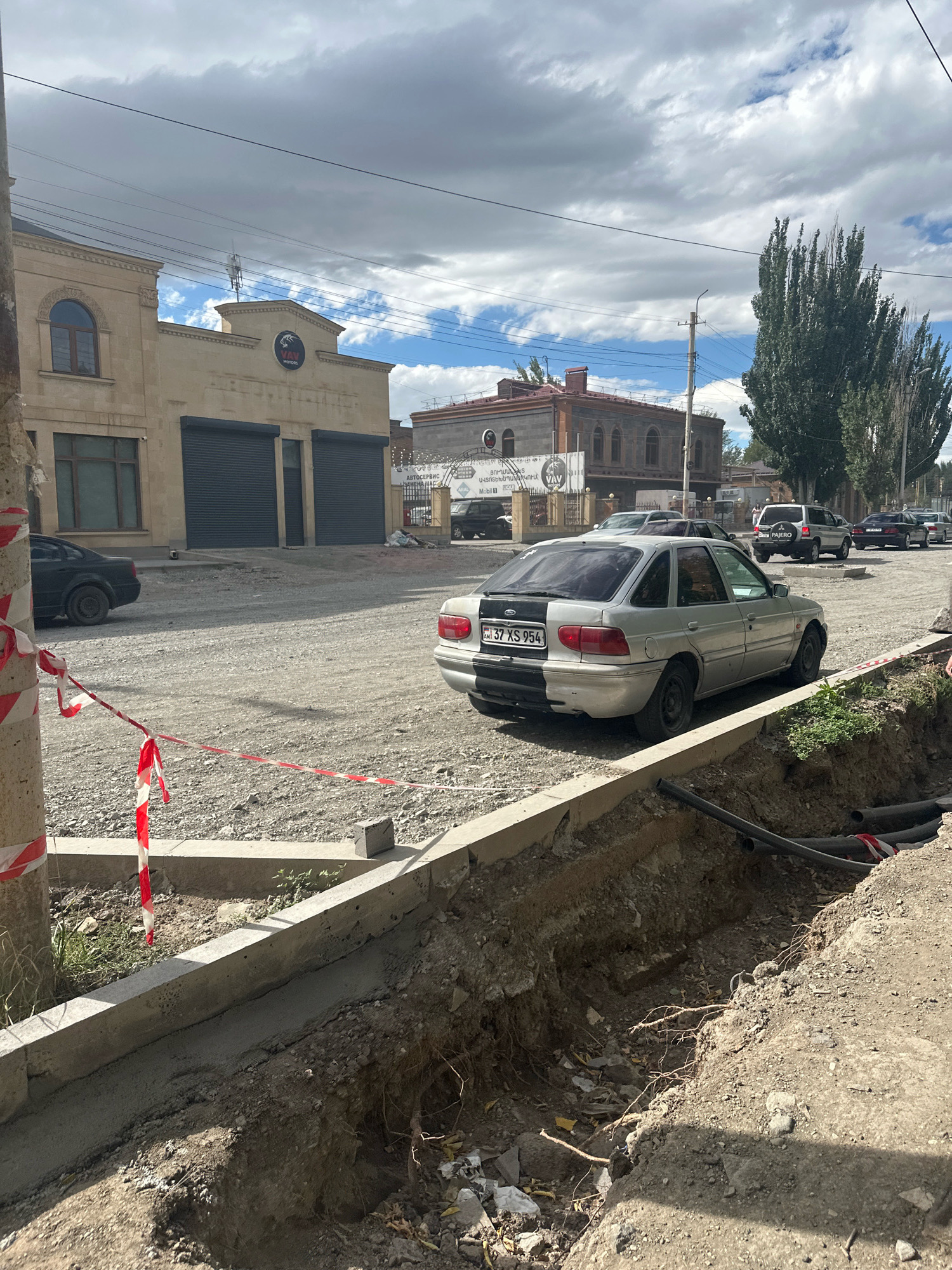 The image depicts a street scene in Gyumri, Armenia, with construction work in progress. A trench has been dug along the side of the road, with concrete curbing being laid. Construction tape, red and white, is visible on a pole and across the area. A silver sedan with a black and silver split paint job on its rear is parked on the gravel road. In the background, several buildings are visible, including one with large grey garage doors and another with arched windows. Trees line the right side of the street, and a Mitsubishi Pajero SUV is also parked on the road. The sky is partly cloudy, with sunlight casting shadows on the ground. The overall impression is one of ongoing urban development and maintenance.