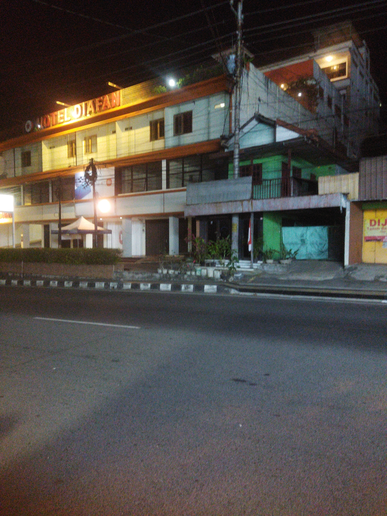 The image depicts a nighttime street view in front of a building identified by a sign reading "HOTEL DIAFAN" in illuminated orange lettering. The building appears to be a hotel or a commercial establishment with multiple stories. The facade features windows, balconies, and a green-colored section on the lower level. In front of the building, there's a sidewalk with some greenery and a tent-like structure. A street with white lane markings is visible in the foreground. The overall scene is illuminated by artificial lighting, indicating it is nighttime. The context provided places this scene in the city of Wonogiri, Indonesia.