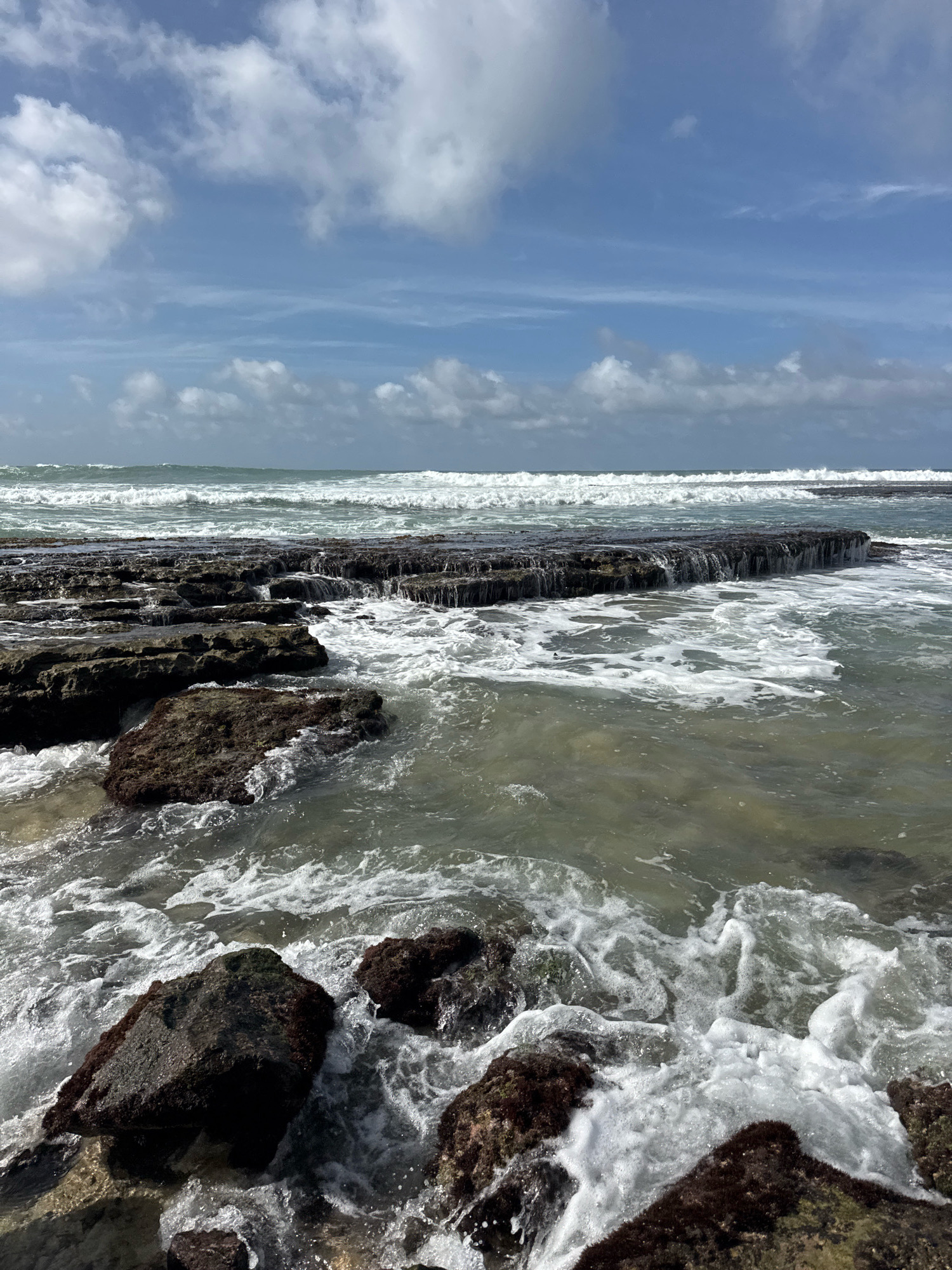 The image depicts a rocky coastline at Goviyapana, Sri Lanka. The foreground is dominated by dark, wet rocks covered in seaweed, with waves crashing and creating white foam around them. In the mid-ground, a series of flat, dark rocks form a natural barrier, with water cascading over their edges. Beyond this, the ocean stretches out, with multiple rows of white-capped waves rolling towards the shore under a partly cloudy blue sky. The water appears murky, with sunlight reflecting off its surface in places. There are no people or man-made structures visible in the image. The scene is natural and dynamic, showcasing the power of the ocean against the rocky landscape.