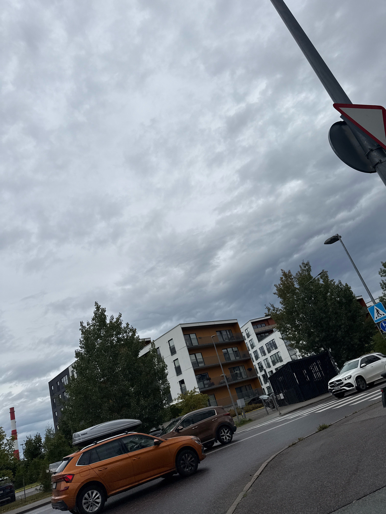 The image depicts a cloudy day in Tallinn, Estonia. In the foreground, an orange SUV with a Thule roof box is parked on the right side of a wet asphalt road. To the right of the orange SUV, a brown SUV is also parked. Further to the right, a white Mercedes-Benz SUV is stopped at a pedestrian crossing. In the background, modern apartment buildings with dark facades and balconies are visible. A tall, slender lamp post with a traffic sign on it stands in the upper right portion of the image. A pedestrian crossing sign with a blue background and a white silhouette of a person walking is also present. To the far left, a red and white striped chimney rises into the sky. The sky is overcast with heavy grey clouds.