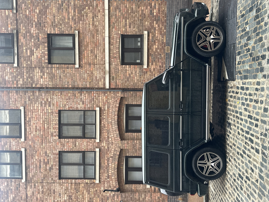 A dark green Mercedes-Benz G-Class SUV, featuring multi-spoke alloy wheels with red brake calipers, is parked on a wet, grey cobblestone street. The vehicle is oriented in profile, facing right, with its front right tire resting on a black rubber speed bump. The background consists of a multi-story building constructed from reddish-brown brick. The building features several dark-framed, recessed windows; some on the lower levels are fitted with horizontal blinds, while others show dark interiors or sheer white curtains. Arched brick lintels are visible above some ground-floor windows. A grey vertical downpipe is affixed to the brickwork on the right side of the building. No human subjects are present. This scene is located in the city of London, United Kingdom.