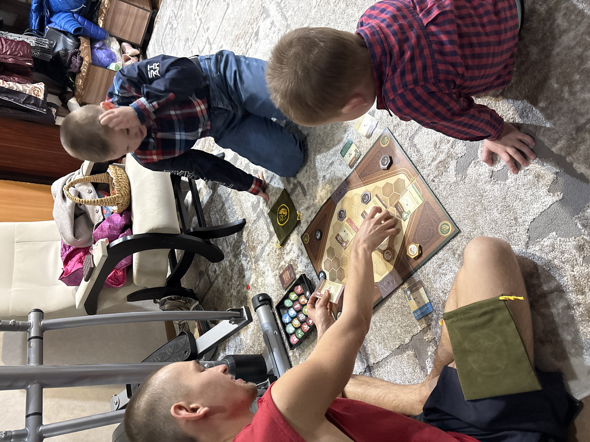 Three individuals are engaged in playing a board game on a patterned rug indoors. Two children, one in a blue and red plaid shirt and jeans, and the other in a red and blue plaid shirt, are positioned on their knees around the board. An adult, wearing a red shirt and dark shorts, is seated with the game board in front of them, holding game cards. The game board is hexagonal and features various illustrations. Several game components such as cards, tokens, and a carrying case are scattered around the board. In the background, a white chair, an exercise machine, and storage containers with assorted items are visible. The location is unidentified, and there's no indication it is in Ulyanovsk, Russia.