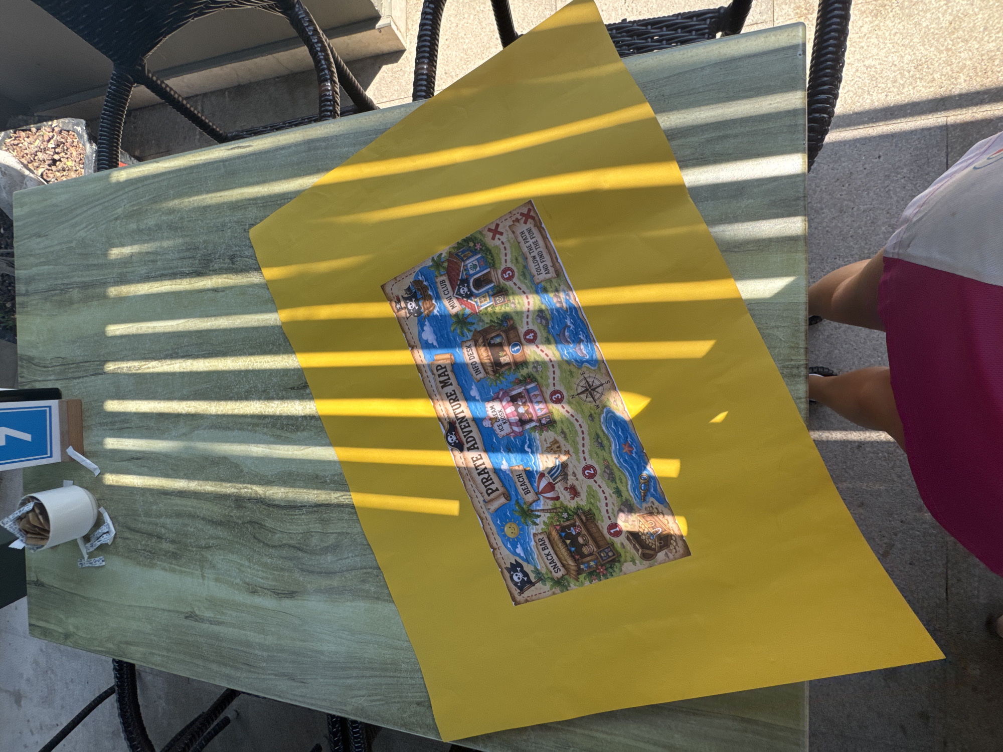 A high-angle, medium shot shows a yellow sheet of paper with a pirate-themed map on a light green table outdoors. The map displays a cartoonish treasure island with various locations such as "SNACK BAR", "BEACH", "ICE CREAM KIOSK", "INFO DESK", and "MINI CLUB." The map is laid out at an angle, covering a portion of the table. Sunlight casts strong diagonal shadows across the scene, suggesting it is daytime. In the background, chairs with dark woven backs are visible, along with a white cup filled with sugar packets and a table number "7" card on the table. A person's legs and lower torso, wearing a pink and white garment, are visible at the bottom of the frame. The ground appears to be paved with stone or concrete. The scene is likely an outdoor seating area of a restaurant or cafe. The city of Aksu, Turkey is not depicted in the image.