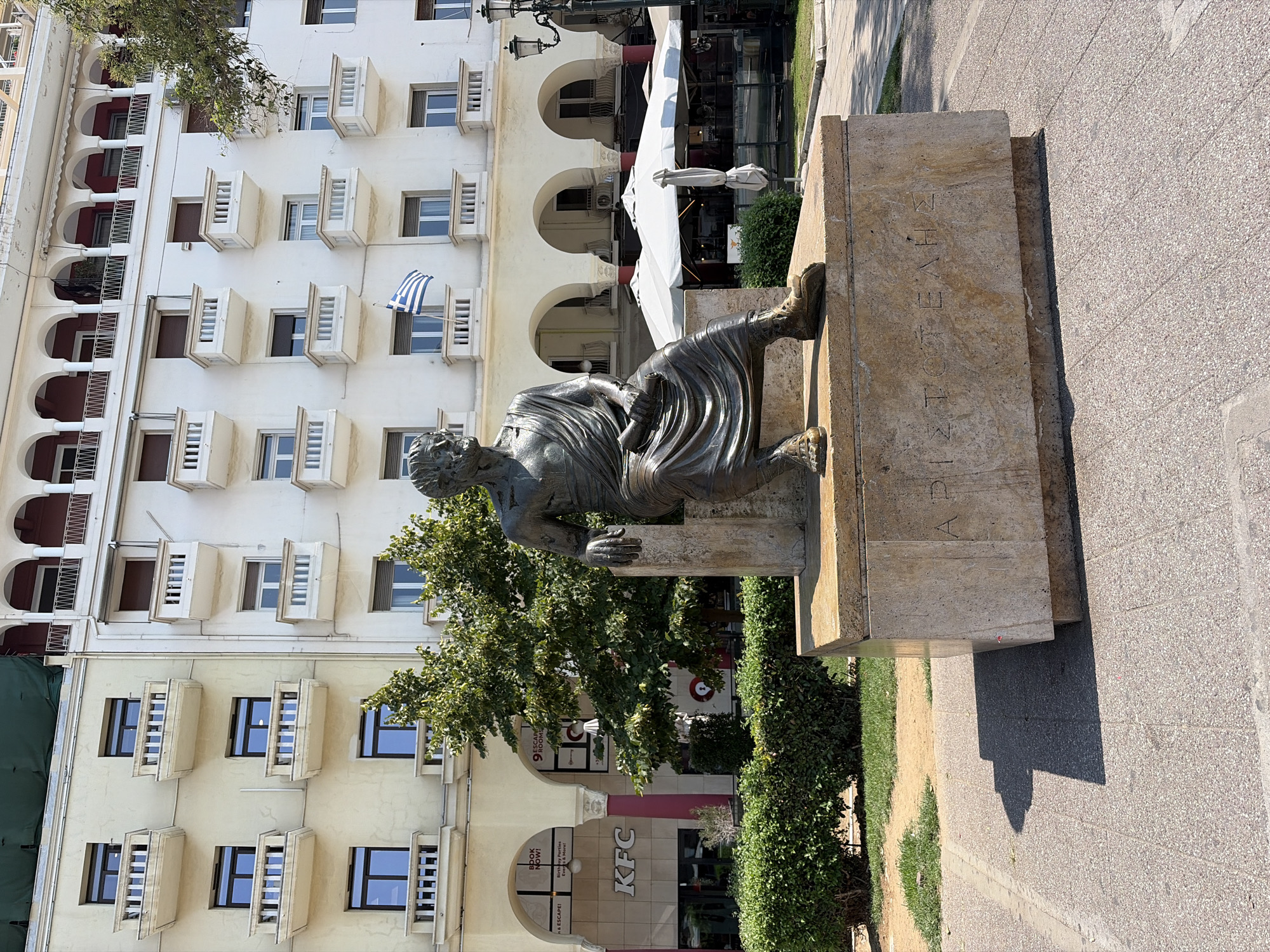 The image depicts a bronze statue of Aristotle seated on a stone pedestal in Aristotelous Square in Thessaloniki, Greece. The statue is positioned in the foreground, with its gaze directed slightly to the left. The pedestal features engraved Greek text which reads "ΑΡΙΣΤΟΤΕΛΗΣ" (Aristotle). Behind the statue, a multi-story building with arched colonnades on the ground floor is visible. Several balconies with white railings are present on the upper floors of the building. A Greek flag is flying from one of the balconies. Green trees and bushes are planted in front of the building, and outdoor seating with umbrellas suggests a cafe or restaurant area. The ground is paved with textured tiles. The overall scene suggests a sunny day in a public urban space.