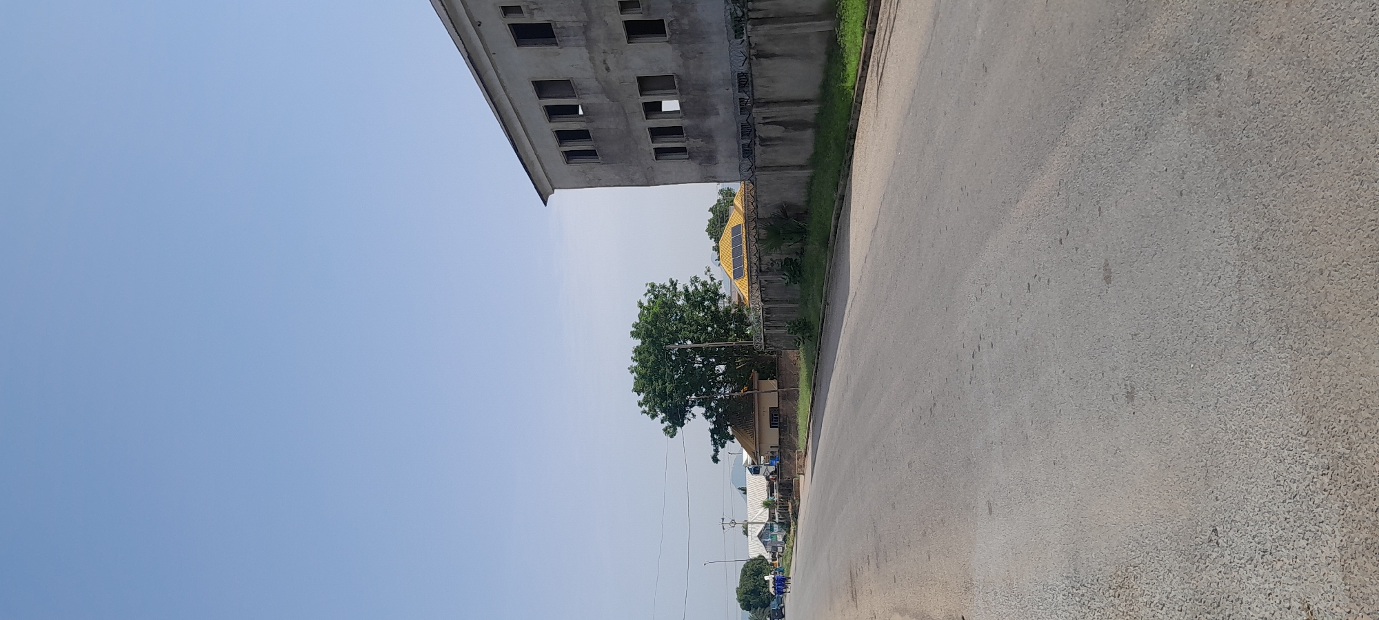 The image shows a street scene in Jalingo, Nigeria. The street is paved with asphalt and appears to be a two-lane road. On the right side of the road, there is a large, unfinished building with multiple window openings. Adjacent to the building is a concrete wall topped with barbed wire. Further down the street on the left, there are several smaller buildings, some with corrugated roofs, and a large green tree. In the distance, there are hills. A few people, dressed in blue uniforms, are walking on the left side of the street. The sky is clear and blue.