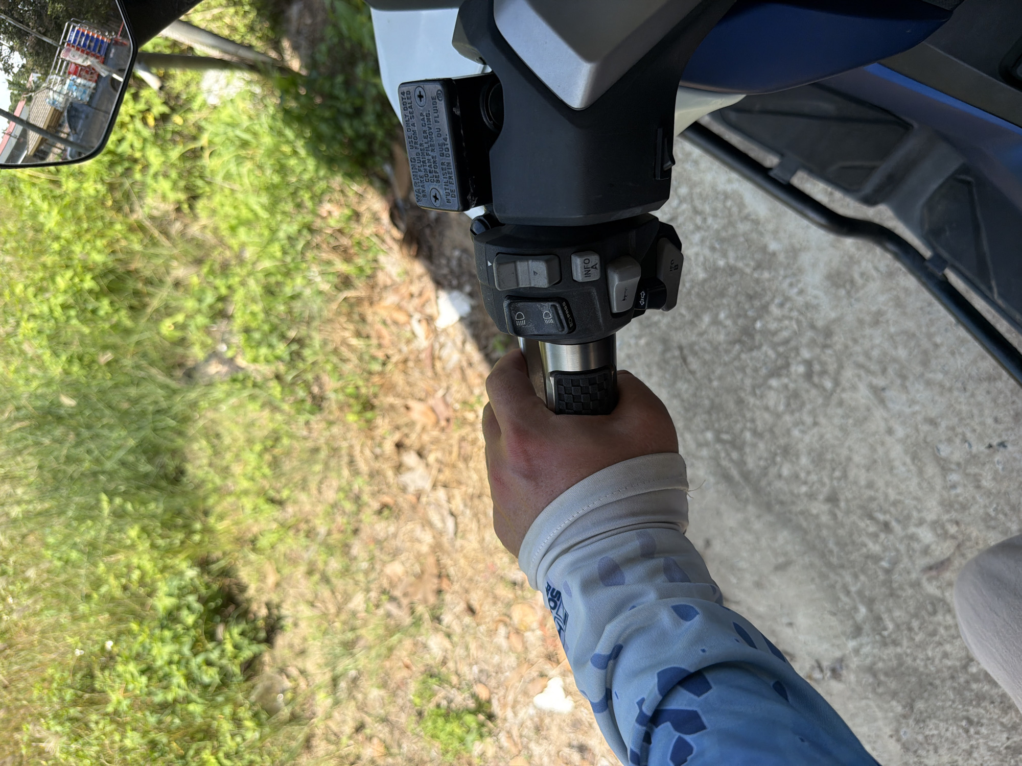 The image shows a close-up view of a person's hand and arm on a motorcycle handlebar. The hand is gloved and gripping the handlebar, which has several buttons and a brake fluid reservoir. The background is out of focus but appears to be a natural setting with greenery and dry ground. A mirror on the handlebar reflects a street scene with buildings and vehicles. The setting is Baan Bo Phut, Thailand.