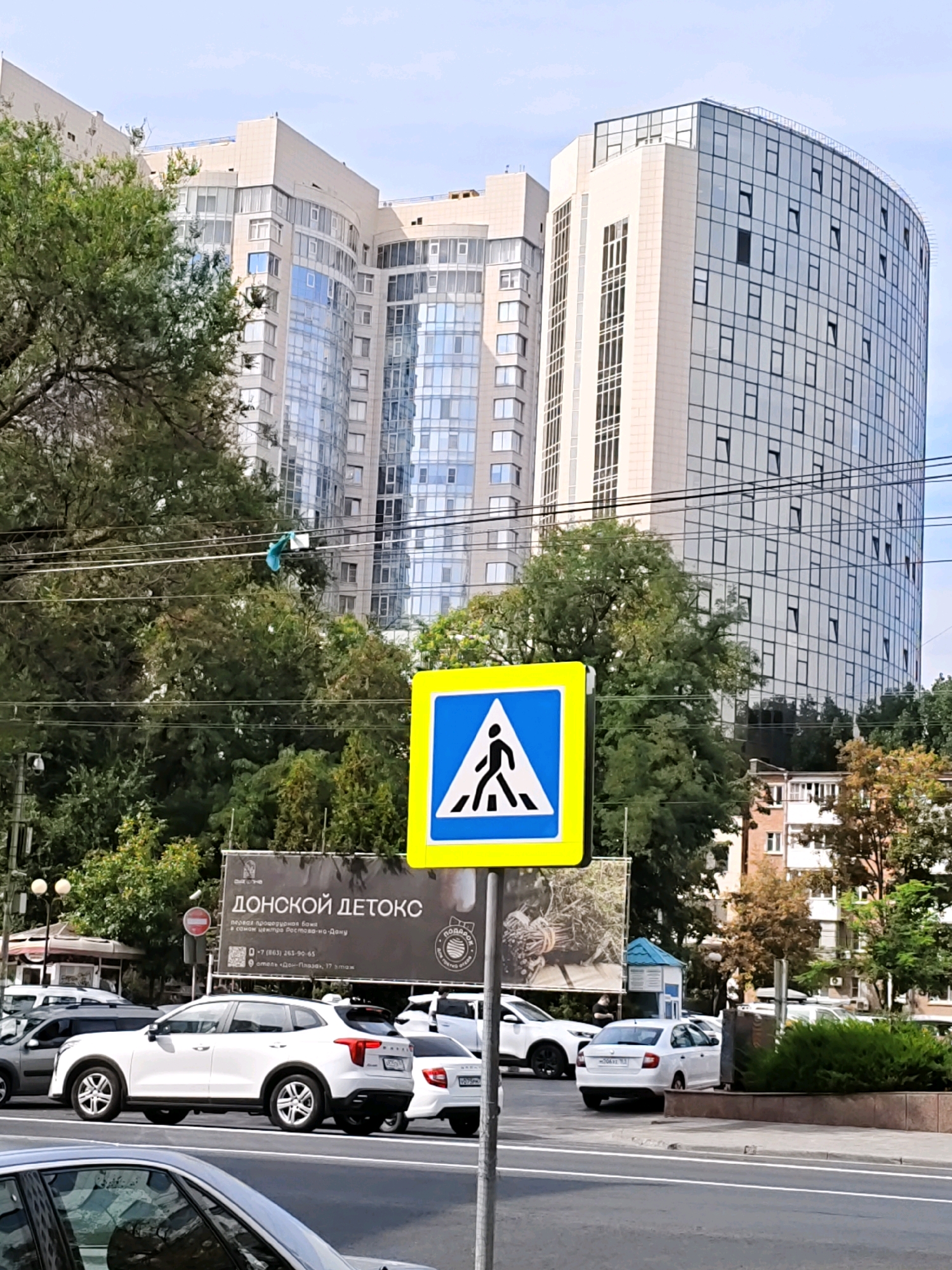The image depicts a street scene in Rostov-on-Don, Russia, on a clear day. In the foreground, a pedestrian crossing sign with a blue background and a white pedestrian silhouette is prominent. Several cars are visible on the road, including a white SUV in the center, a white sedan behind it, and parts of other vehicles. The background features modern, multi-story buildings with large glass facades, typical of an urban environment. Lush green trees line the street, partially obscuring some of the buildings. A billboard advertisement for "ДОНСКОЙ ДЕТОКС" (Donetsk Detox) is also visible. Power lines are strung across the scene. The overall impression is of a busy urban thoroughfare with significant architectural elements and everyday city life.