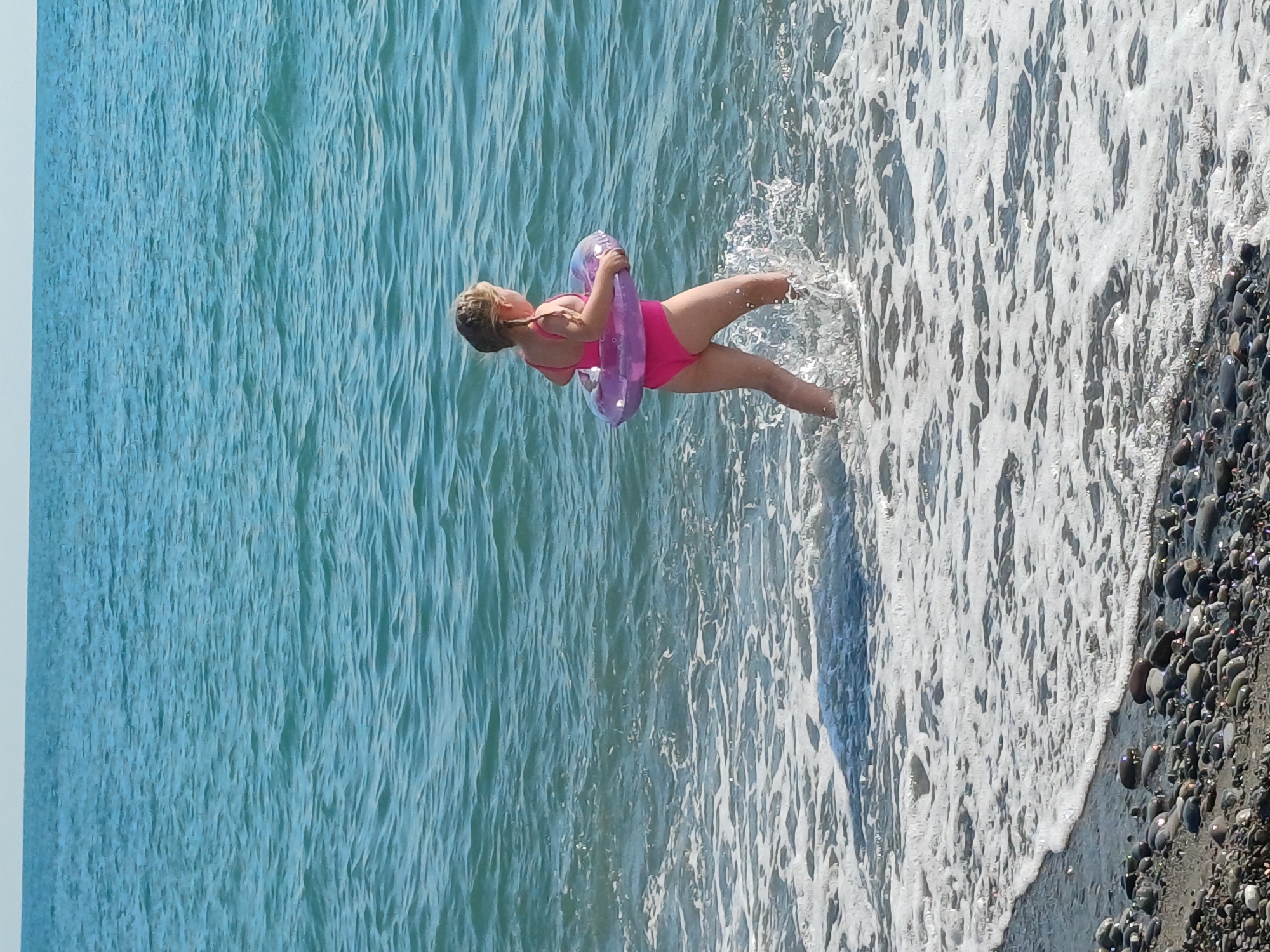 A young girl in a pink swimsuit stands in the shallow water at the edge of a beach, holding a purple inflatable ring. She is facing left, with her body angled towards the water. The water is a clear, turquoise blue, with gentle waves lapping at the shore. White foam from the waves spreads across the pebbles on the beach. The beach is made up of dark, smooth pebbles. The sun is shining, creating bright highlights on the water and the girl's skin. The scene appears to be a sunny day at the seaside, possibly in Sochi, Russia, given the rocky shoreline. The girl's pose suggests she is about to enter the water or is enjoying playing in the shallow waves.