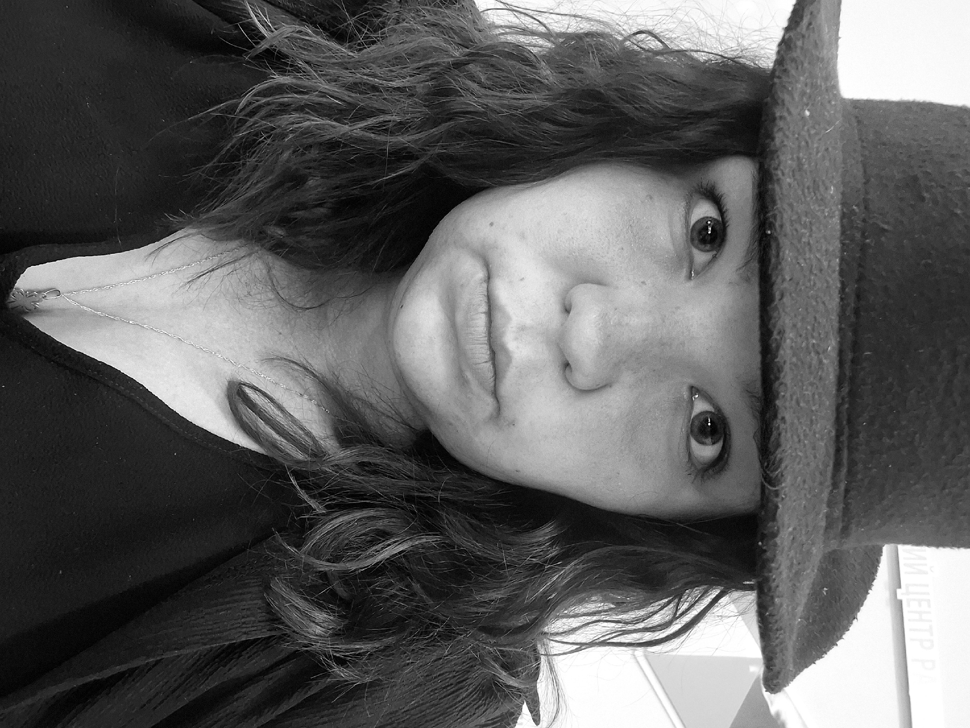 A grayscale close-up shows a woman with wavy hair, wearing a dark top and a hat, with a necklace visible around her neck. Text, partly obscured, appears to read "МИ ЦЕНТР Р..." in the bottom right corner.