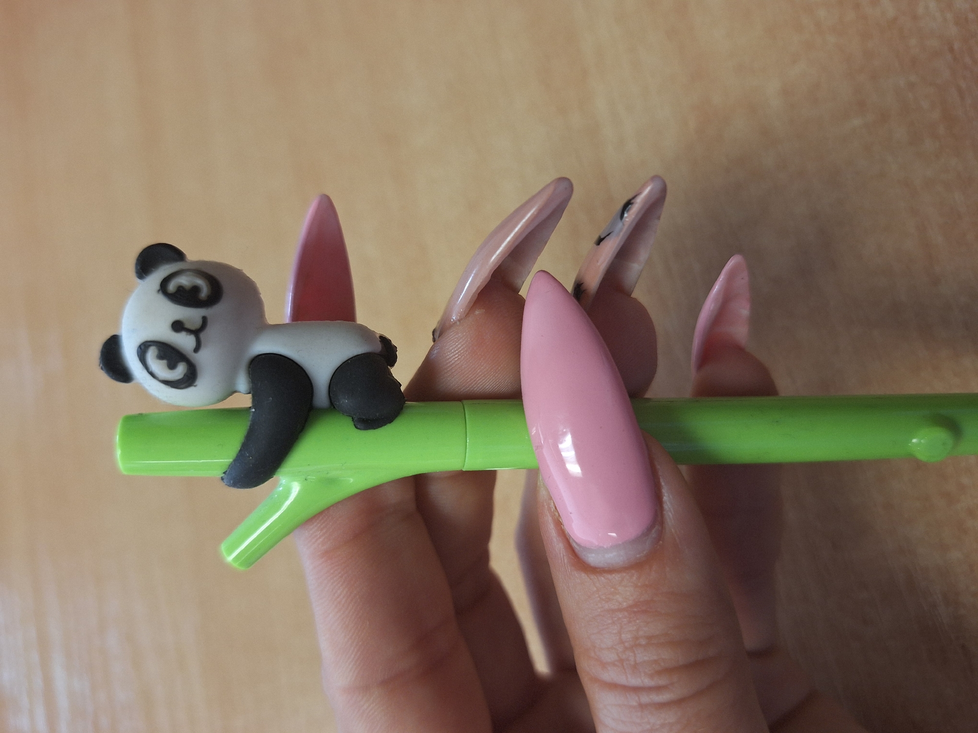 A hand is holding a green plastic pen with a toy panda clinging to it. The hand has long, pointed artificial nails, painted pink and nude colors. The background is a light brown, possibly a wooden surface. The location is unknown.