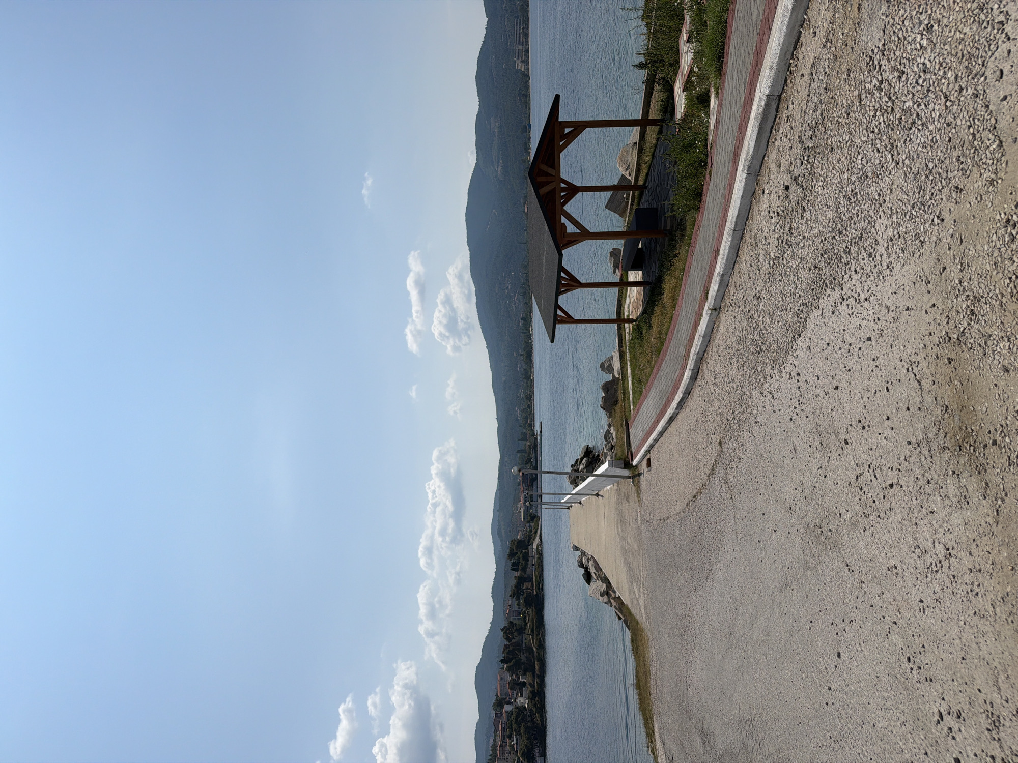 The image depicts a scenic view of the coastal area within the Sithonia Municipal Unit, Greece. In the foreground, a paved pathway, composed of rough gravel, curves gently to the right, bordered by a red brick pathway and a concrete curb. Further ahead, a concrete pier extends into a calm, blue body of water. Along the pier, lampposts are spaced at intervals, and a low white railing lines its right side. The water is surrounded by a hilly coastline with scattered buildings and lush green vegetation. In the middle ground, to the right of the pier, a wooden gazebo with a dark roof and benches underneath is situated on a stone-tiled area, surrounded by rocks and grass. The sky is a clear, bright blue with scattered white cumulus clouds. The overall scene conveys a peaceful and sunny day at the seaside.