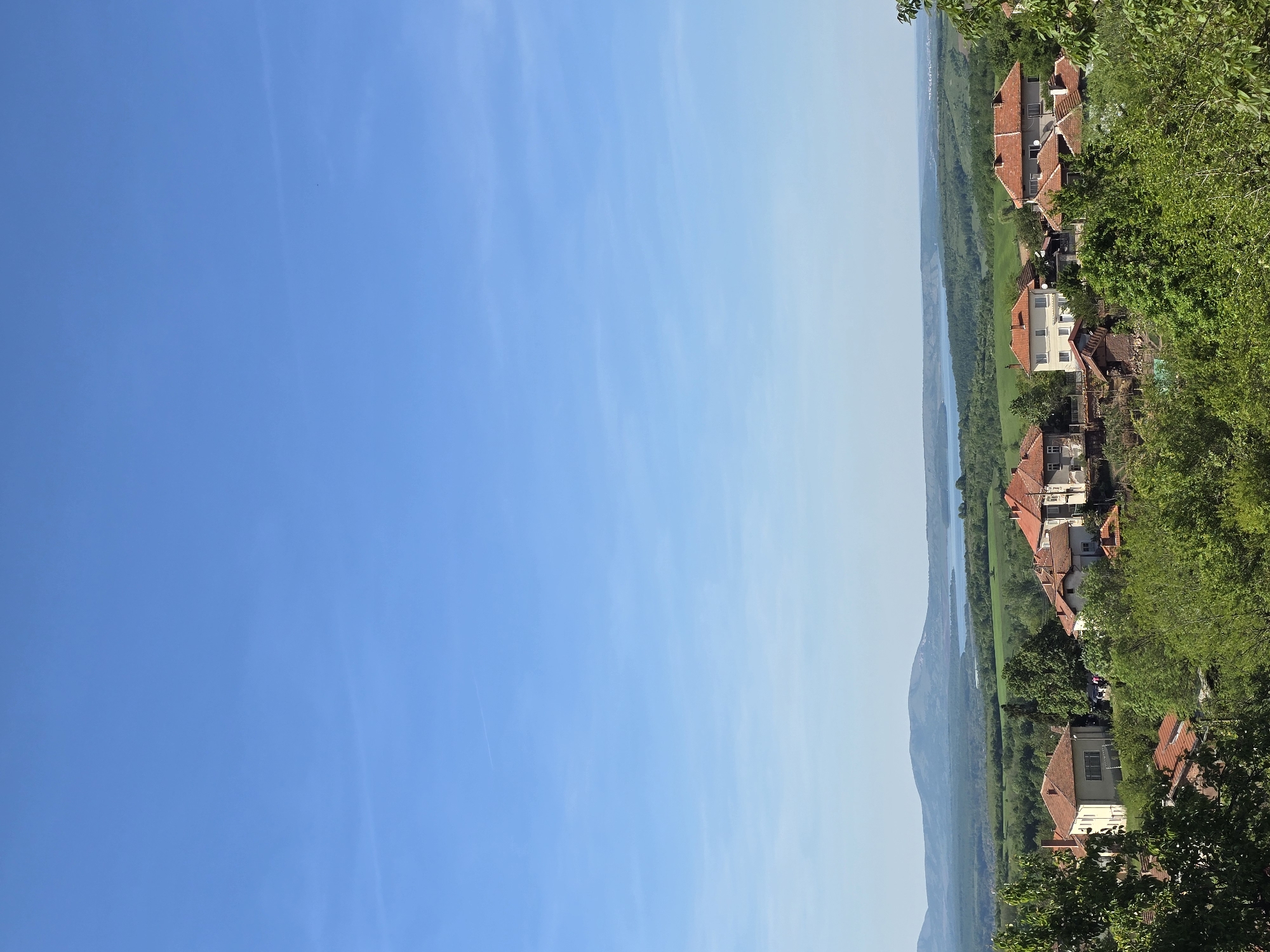The image shows a view of Bokilovtsi, Bulgaria. The right side of the image depicts houses with red tile roofs nestled amidst lush green trees. In the distance, a river flows through a valley with a mountain visible in the background. The left side of the image is dominated by a clear blue sky.