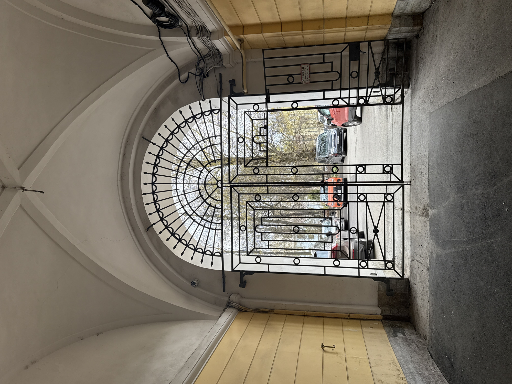 The image, initially rotated 90 degrees clockwise, depicts a view looking outwards through an arched passageway. The interior of the archway features a painted, light-colored vaulted ceiling with visible cracks and numerous black electrical cables running along its contours, secured with clips. Two small, white surveillance cameras are mounted near the top of the arch. On the right side of the passageway, a section of ceiling is covered with light yellow tiles, and a yellow wooden door with a metal handle is visible. The floor transitions from this yellow paneling to a darker, rough concrete surface towards the left.
The archway is closed off by an elaborate, black wrought iron gate. The top portion of the gate mirrors the semi-circular arch, featuring radiating bars with decorative finials. The lower, rectangular section of the gate has a geometric pattern of horizontal and vertical bars interspersed with circular motifs. A smaller pedestrian gate, or "kalitka," is integrated into the larger structure.
Through the gate, an outdoor scene is visible under what appears to be overcast daylight. Several cars are parked on a grey concrete or asphalt surface, including vehicles in red, gray, and orange/brown hues. Beyond the parked cars, trees with bare branches or very sparse, light-green leaves suggest either late winter or early spring.
A small, red-and-white sign is affixed to the upper right section of the wrought iron gate. The text, in Russian, reads: "ПРОХОД КАЛИТКИ ЗАПРЕЩЕН," which translates to "Passage through the pedestrian gate is forbidden." The overall impression is of an older building, typical of Saint Petersburg architecture, with modern security and utility installations.