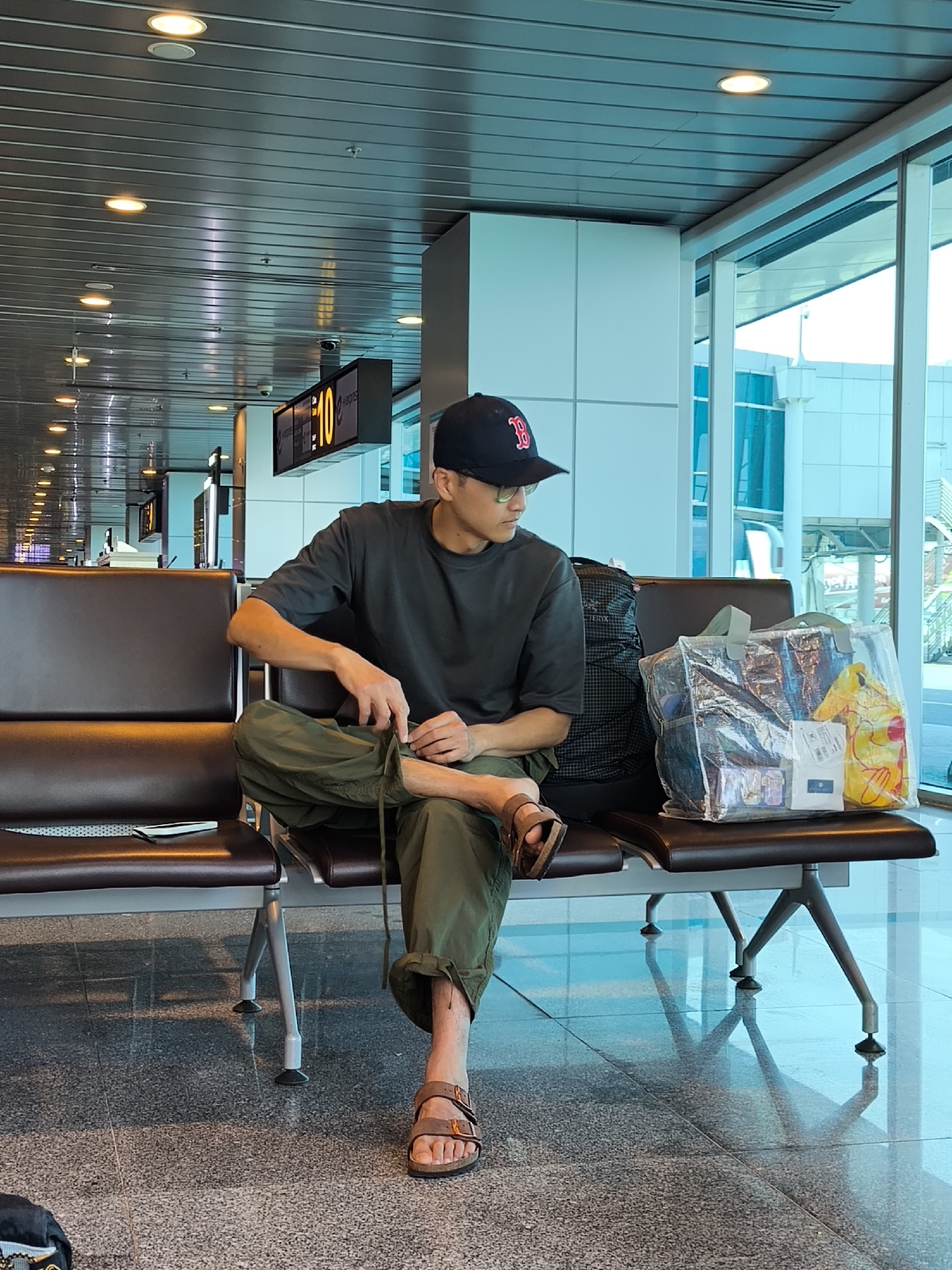 An adult male is seated in a modern airport terminal waiting area, specifically at Gate 10, in the city of Đà Nẵng, Vietnam. He is positioned on a dark brown multi-seat bench with metal legs. The male, wearing a dark grey t-shirt, olive-green cargo pants, and brown sandals, is also sporting eyeglasses and a dark blue baseball cap with a red 'B' logo (Boston Red Sox). His left leg is crossed over his right, and he is actively adjusting a drawstring on his left pant leg with both hands, looking down and to his left.
To his immediate right, on the same bench, rests a black and grey ARC'TERYX backpack and a transparent plastic shopping bag containing a yellow Pikachu-themed item along with other packaged goods. A white smartphone is visible on the seat to his left. The environment features a polished, reflective grey speckled tile floor and a grey slatted ceiling with numerous circular recessed lights. Large windows line the wall to the right, offering a view of the airport tarmac and another building, bathed in natural light. A digital display board indicating "10" for the gate number is visible in the background.