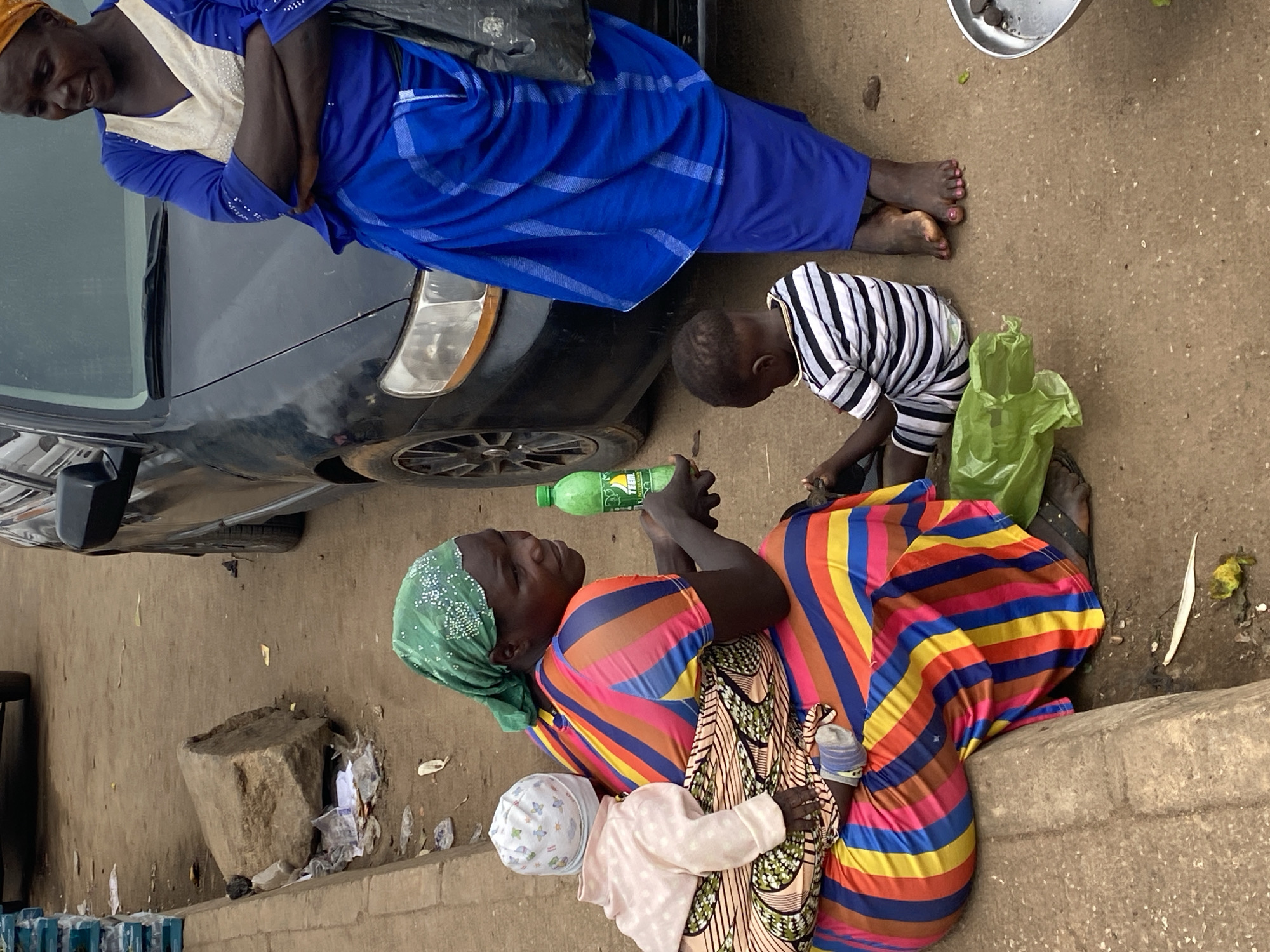 This outdoor scene, set in Bukuru, Nigeria, captures a casual street or roadside gathering during the daytime. The image features four individuals and a parked black car.
On the left side, a woman with dark skin stands barefoot, wearing a blue dress with white detailing on the upper body and a blue head covering. She has a slight smile and her arms are crossed. Her attire suggests traditional or modest clothing. Next to her, a black car is partially visible, parked on what appears to be a dirt or concrete surface.
In the middle of the frame, a young child, also with dark skin, is kneeling or crouching on the ground. The child is wearing a black and white striped t-shirt and is focused on something on the ground, possibly a small object or activity. A bright green plastic bag rests near the child.
On the right, another woman with dark skin sits on the ground, her back against a concrete curb or raised edge. She wears a vibrant, multi-colored striped dress in shades of red, yellow, blue, and green, and a green head covering. She is holding a green plastic bottle with the "Sprite" logo clearly visible. Close to her, an infant or very young child is visible, wearing light-colored clothing and a white hat with a pattern, appearing to be held or sitting very near the woman.
The ground is a mix of dirt and concrete, with some scattered debris including small pieces of paper and what looks like discarded fruit peels. In the upper right corner, a small metallic bowl containing some dark, round objects is visible. A large, rough-hewn stone or block is also on the ground to the bottom left of the sitting woman. The general atmosphere appears calm, suggesting an ordinary moment in daily life.