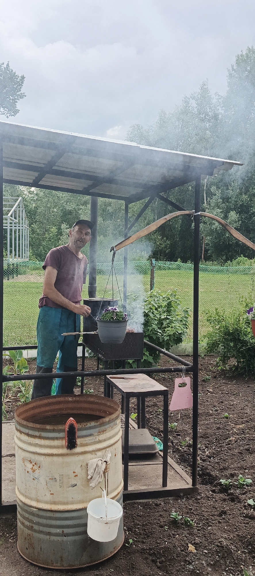 A man wearing a purple t-shirt, turquoise pants, and black boots stands under a metal roofed shelter in a rural outdoor setting. He is looking at the camera and smiling. In front of him is a charcoal grill with smoke rising. A pot with purple and white flowers hangs from a wooden contraption above the grill. To the left of the grill is a large, rusted metal barrel filled with water, with a red and black brush attached to its side. A white bucket hangs from a hook on the barrel. Behind the man is a grassy area with trees in the background and a white fence. The ground in the foreground is dark soil with some small green plants. The scene appears to be in a backyard or garden area, consistent with a location in the city of Chaikovsky, Russia.