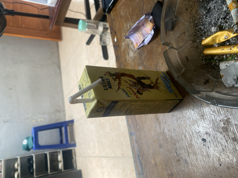 A yellow Ultra Milk carton, with a white flexible straw inserted into its top, rests on a worn, light-brown wooden table. The carton features graphics of a person on a skateboard and the text "Make Your Move!" To the right of the carton, a dark, circular ashtray, patterned with a soccer ball design along its rim, contains ash, several cigarette butts, and two crumpled gold-colored foil wrappers. Adjacent to the ashtray, a crumpled Indonesian Rupiah banknote and a small dark object are visible.
The background shows an indoor environment with light-colored tiled flooring and plain walls. On the left, a multi-tiered shoe rack holds several pairs of shoes. Behind it, a blue plastic stool supports a green baseball cap. On the right, a portion of a dark wooden table is visible, holding a clear bottle with a light green cap, a clear glass, and the base of an electronic fan. A closed wooden door is partially visible in the far right background. No individuals are present. The scene is located in Cimenyan, Indonesia.